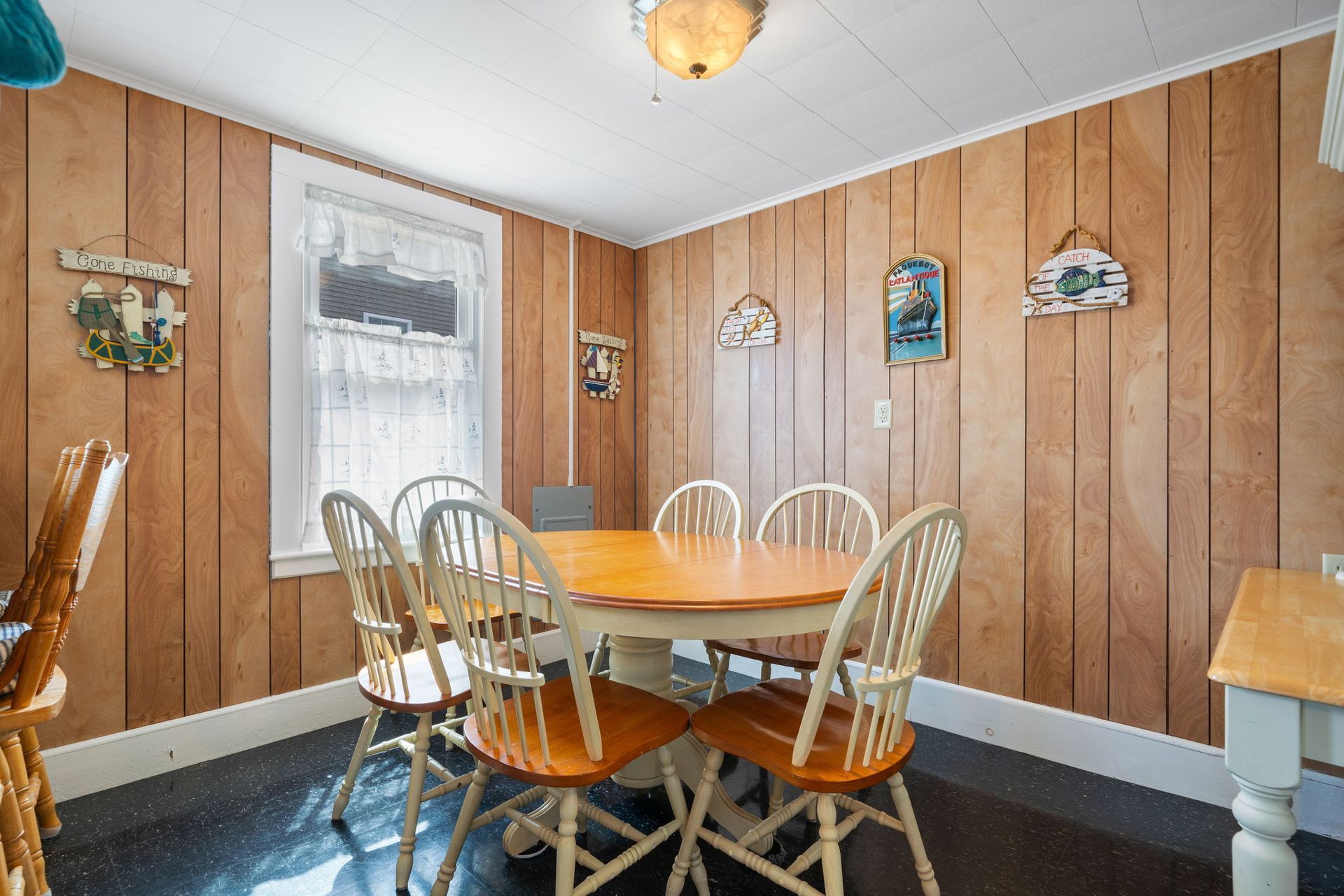 Dining room with round wooden table, six chairs, and wood-paneled walls, window with lace curtain.