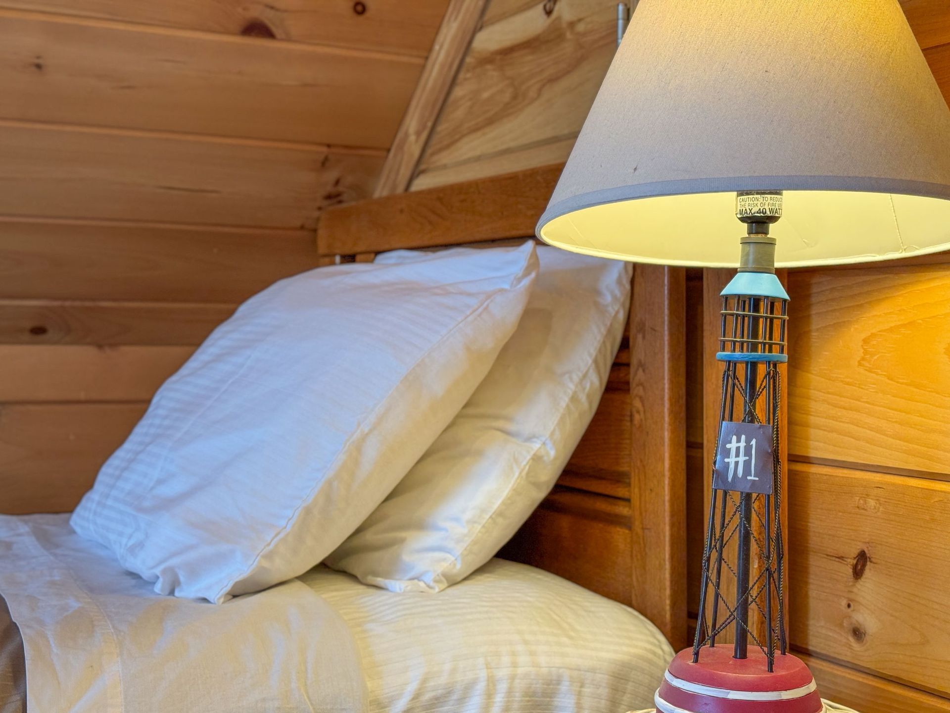 Bed with white pillows, beside a lamp on a wooden nightstand in a wood-paneled room.