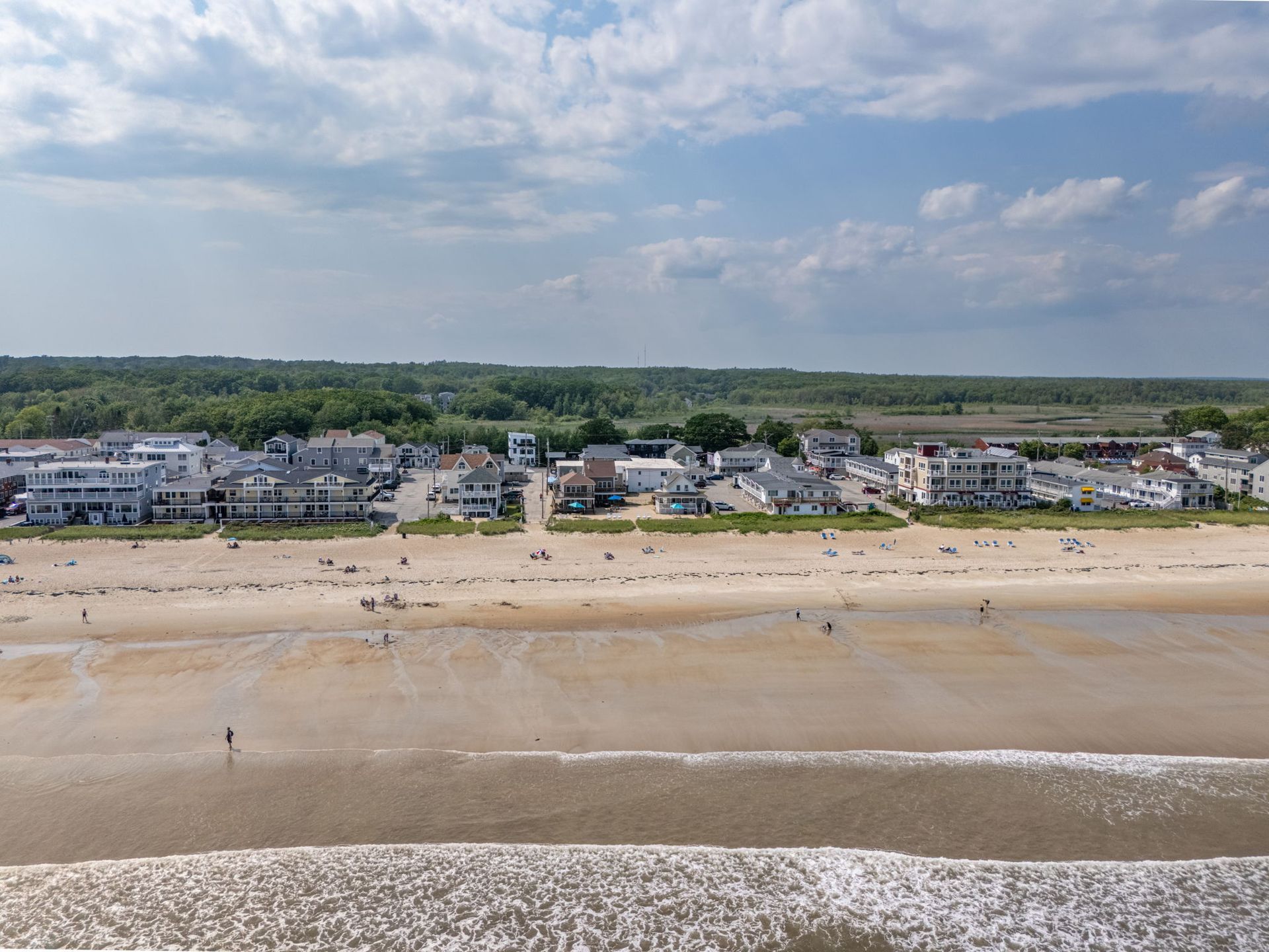 Beachfront with buildings, sand, and ocean under a partly cloudy sky. People are on the beach.