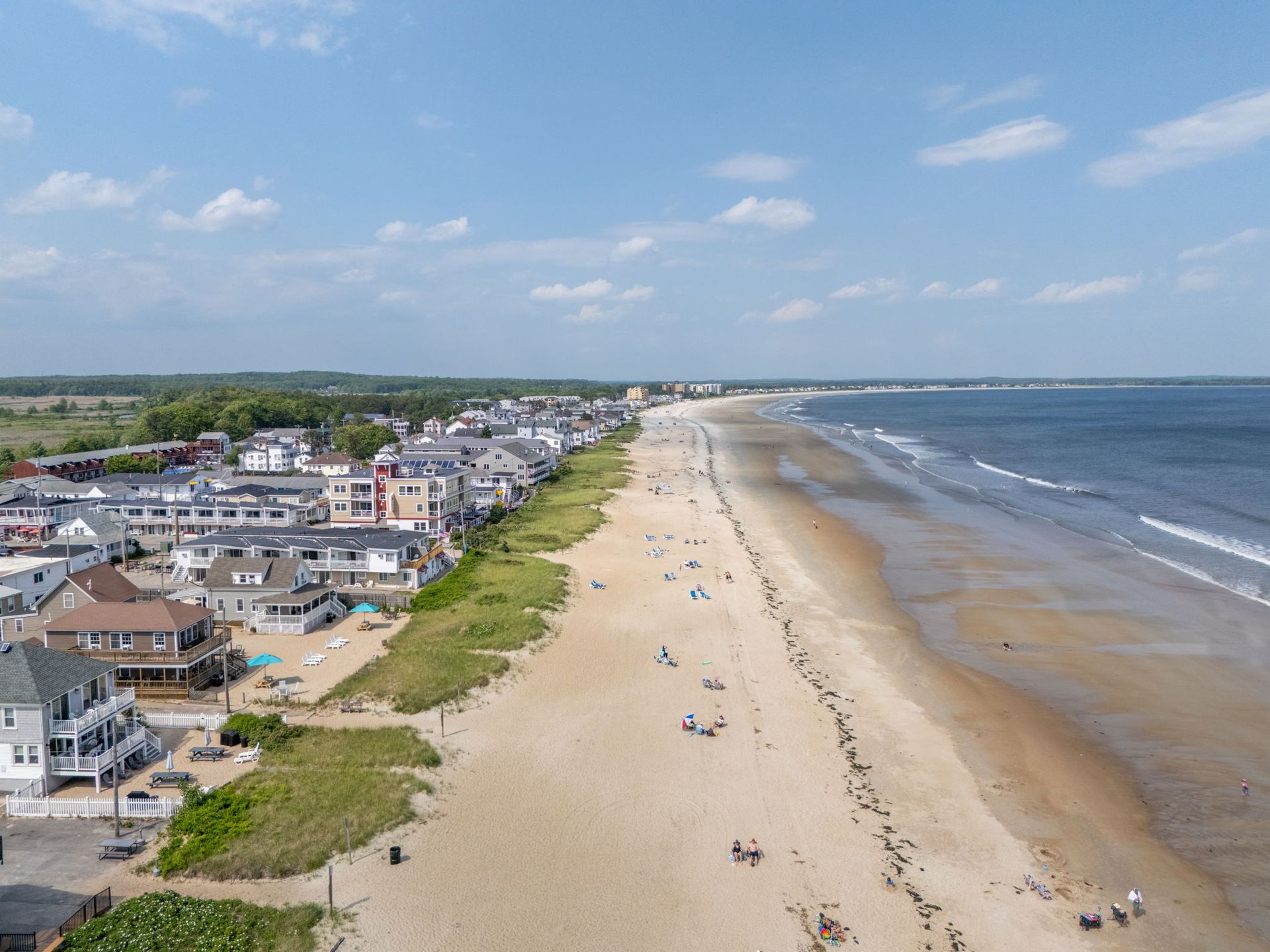Sandy beach lined with buildings under a blue sky. People relax on the beach near the ocean.