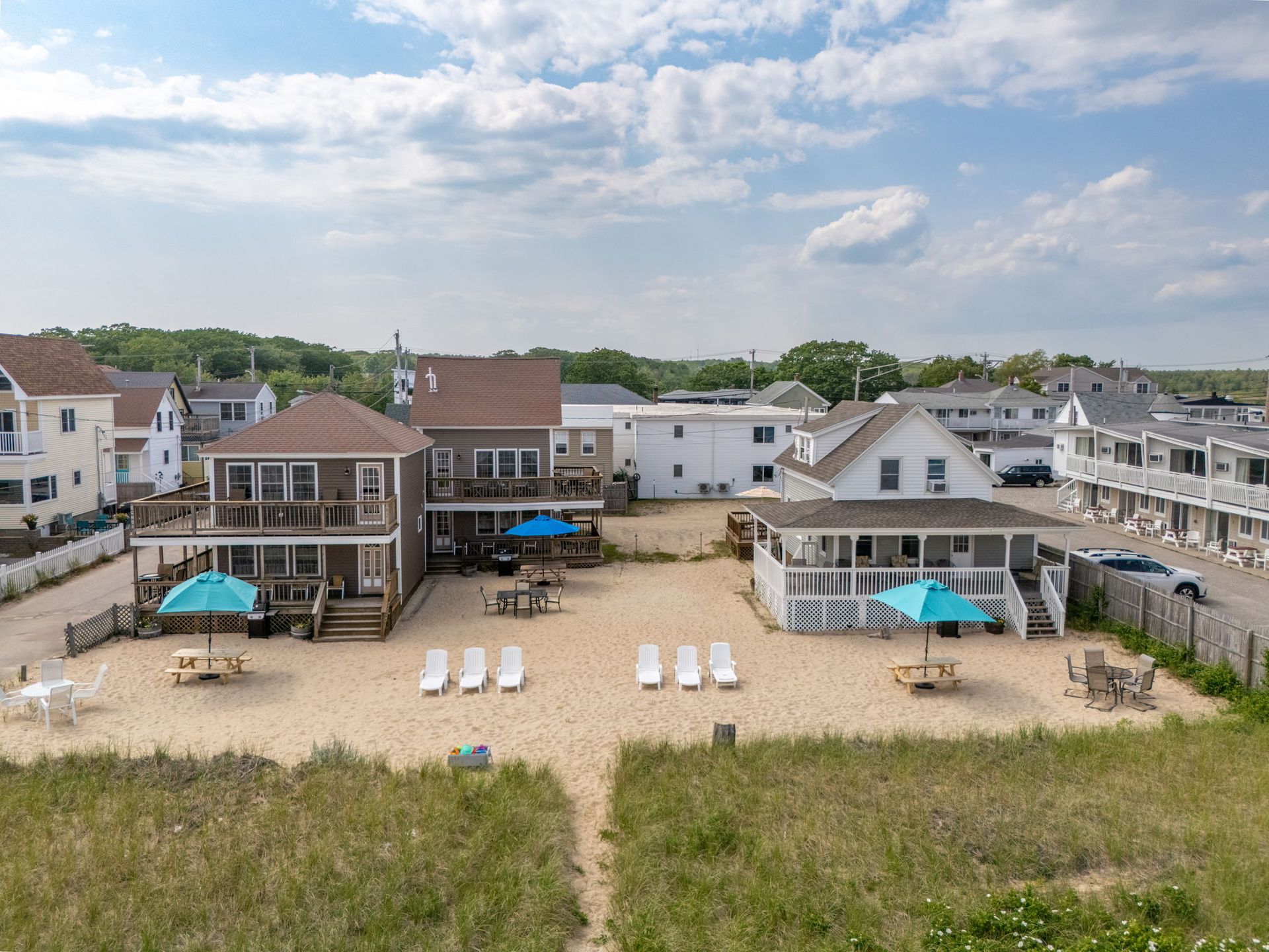 Beachfront homes with decks, sand, umbrellas, and lounge chairs under a blue sky.