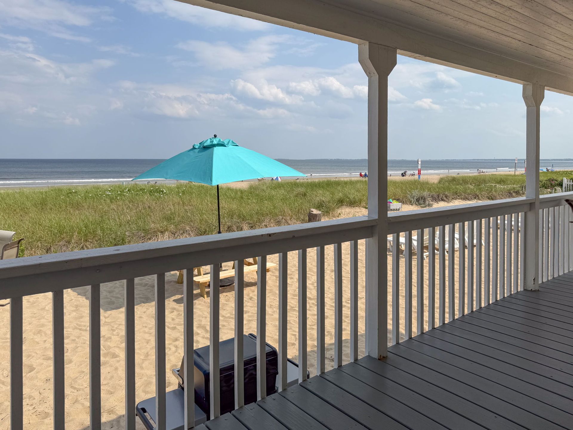 Beach view from a porch. Turquoise umbrella, sandy beach, ocean, blue sky, white railing.