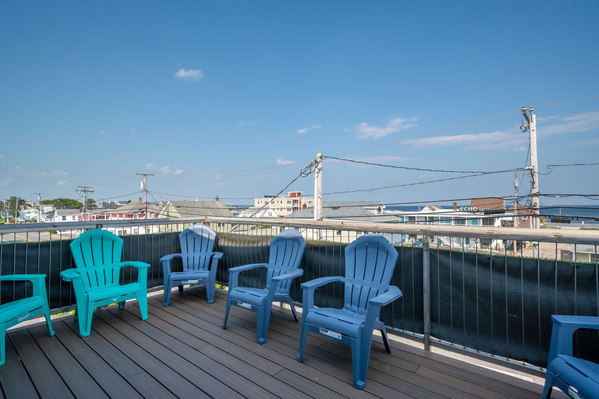 Blue and teal Adirondack chairs on a wooden deck overlooking a coastal town under a blue sky.