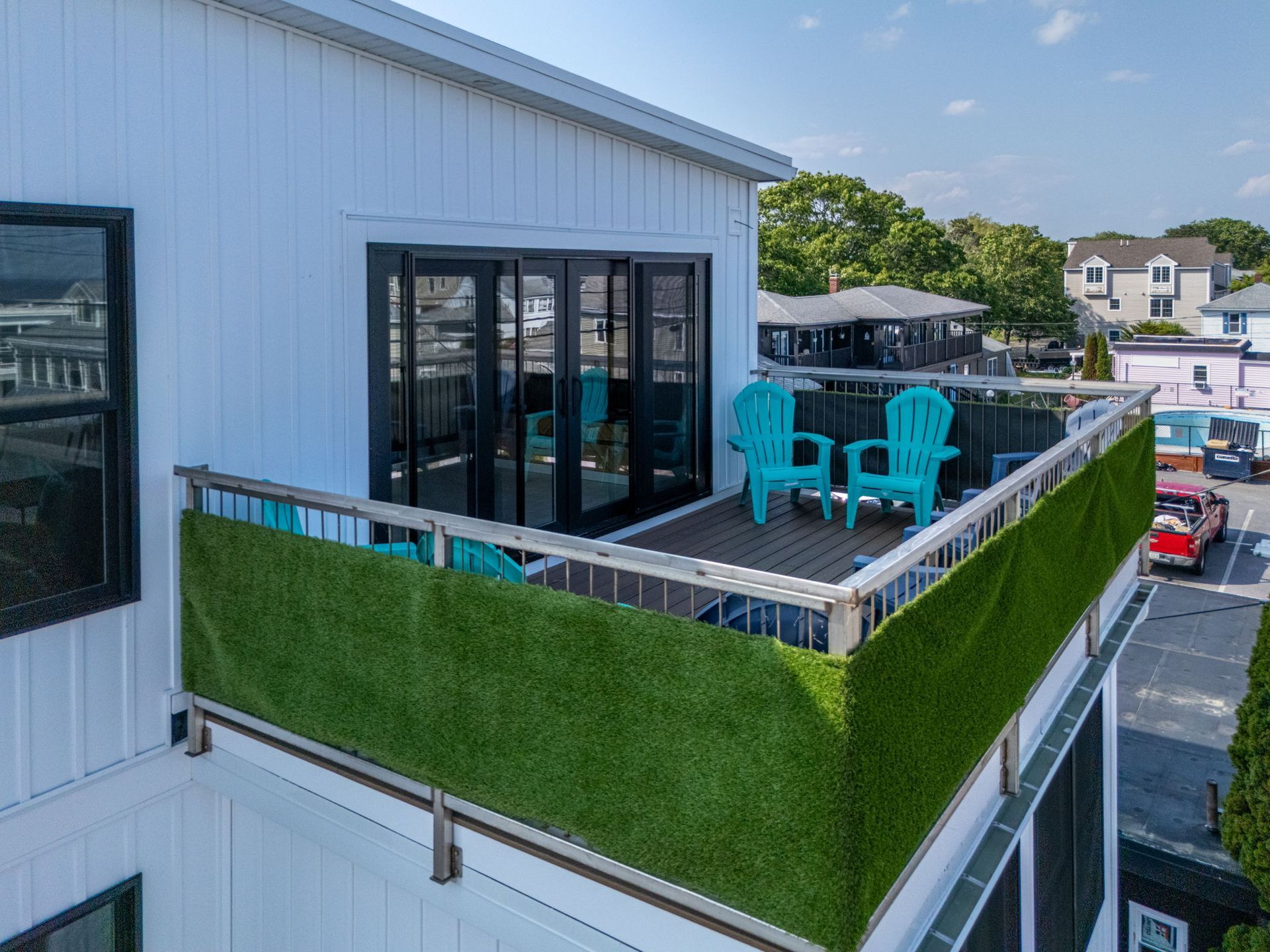 Balcony with green turf barrier, teal chairs, black doors, white building exterior, blue sky.