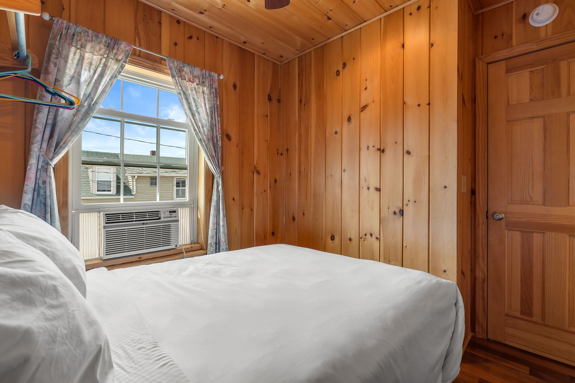 Bedroom with a white bed, window with curtains, and wood-paneled walls. An air conditioner sits in the window.