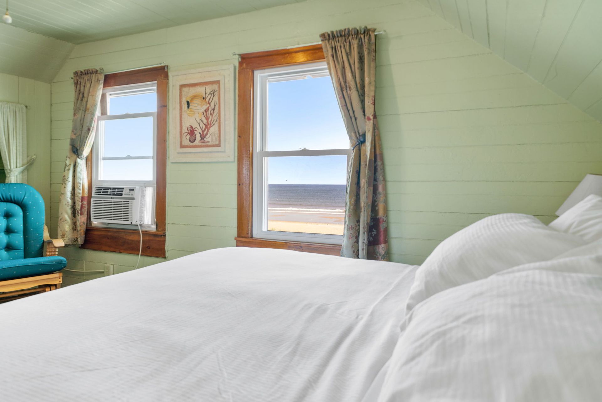 Bedroom with a bed facing a window overlooking the ocean. Green walls and a teal chair.