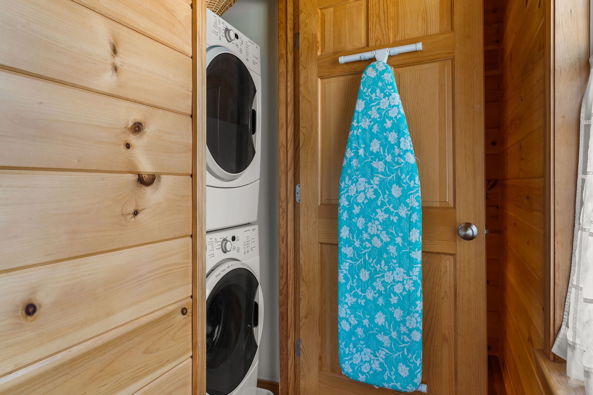 Stacked white washer and dryer in a closet, with a teal ironing board hanging on the door.