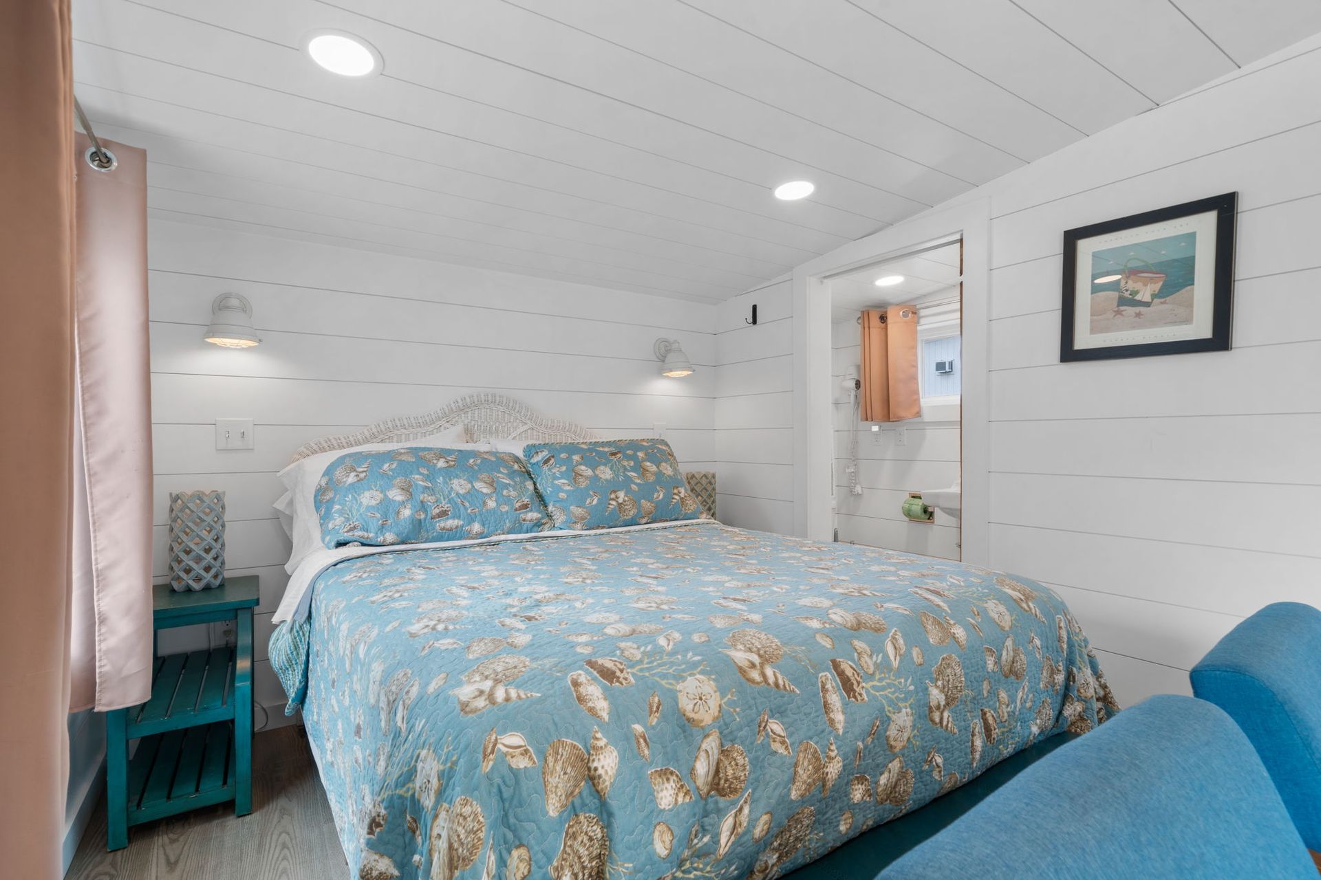 Cozy bedroom with a blue seashell quilt, turquoise nightstand, and a small bathroom visible. White walls.
