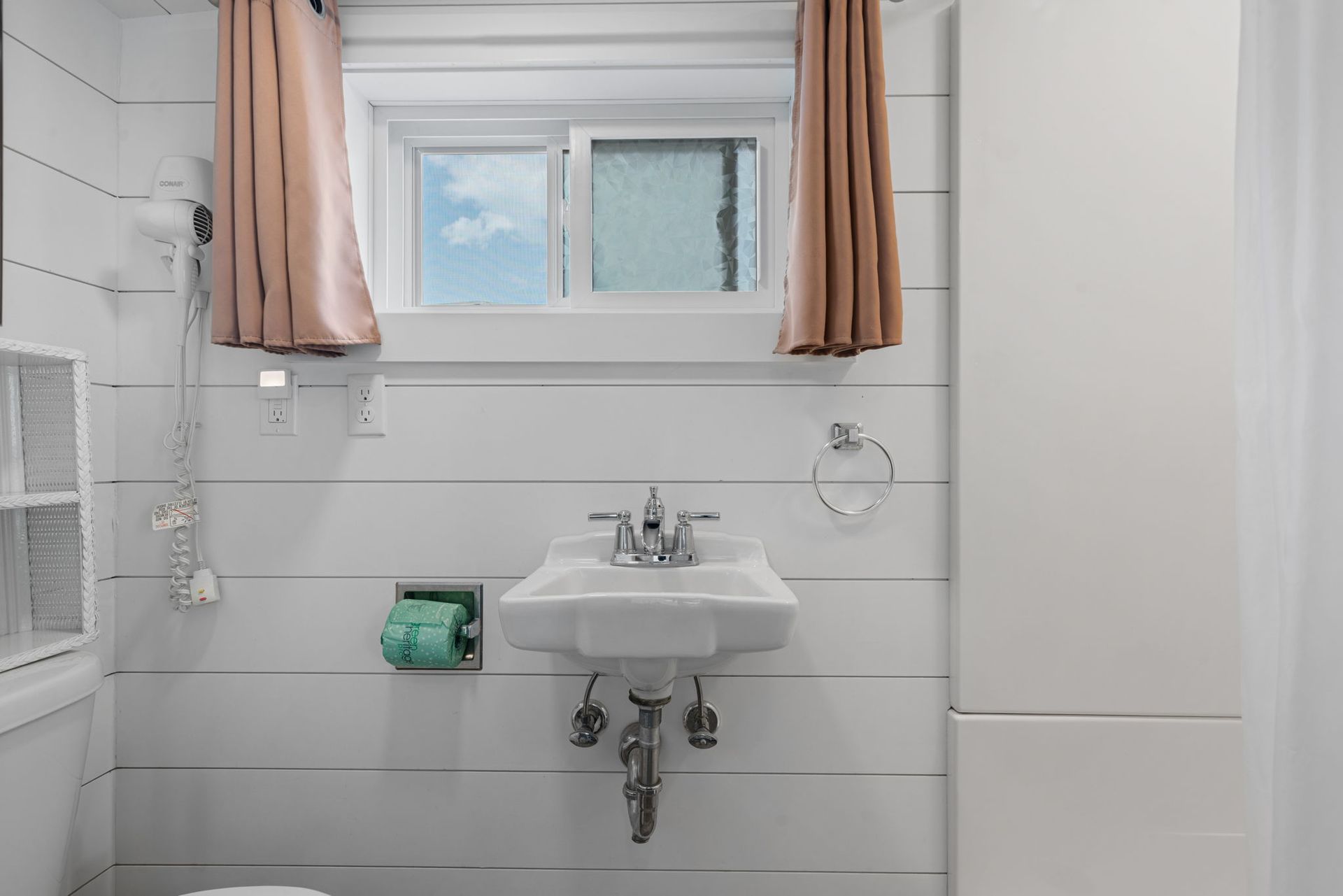 Small, white bathroom with a sink under a window; pink curtains, teal toilet paper holder.