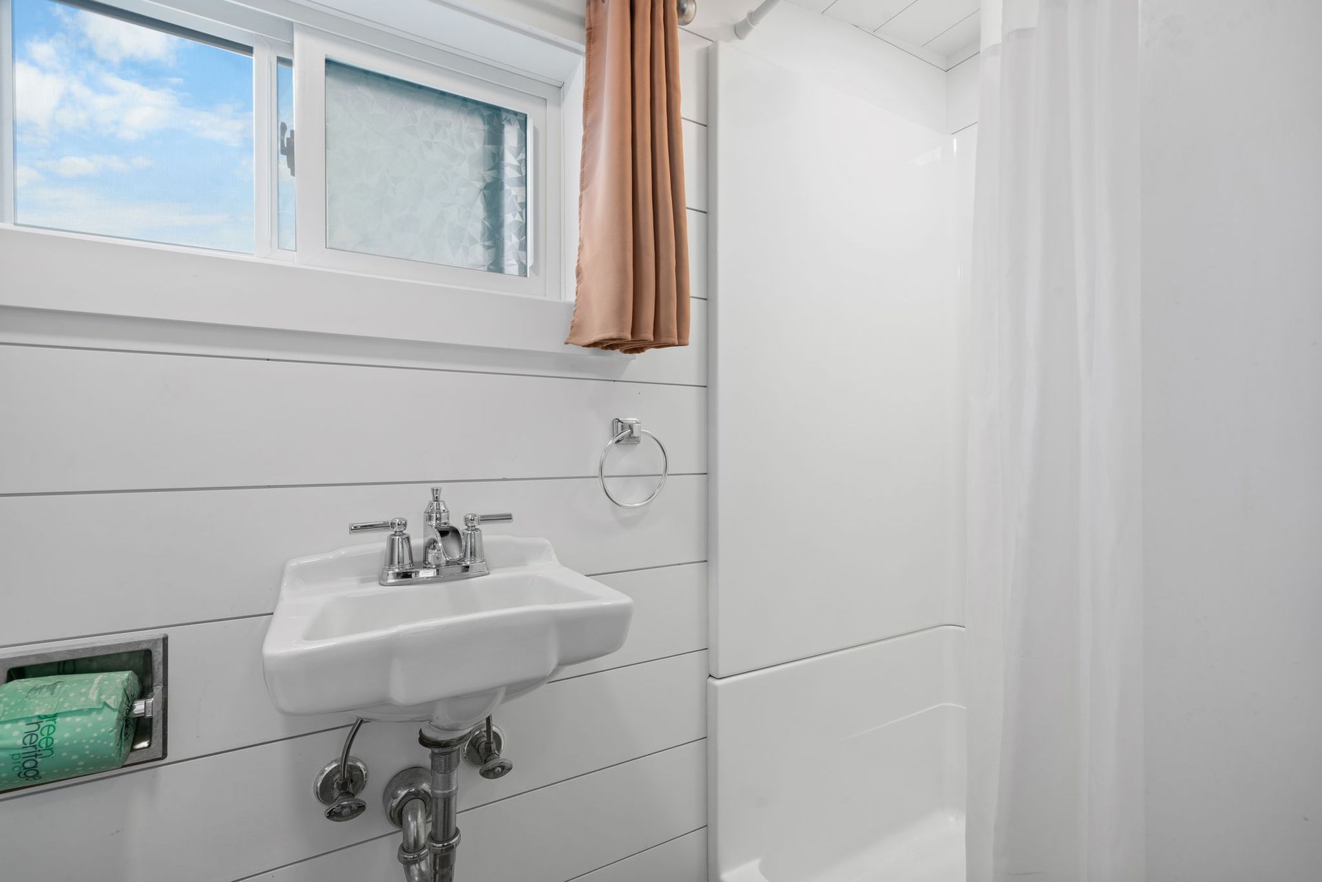 Small bathroom with a window, sink, and shower. White walls, pink curtain, turquoise hand towel.