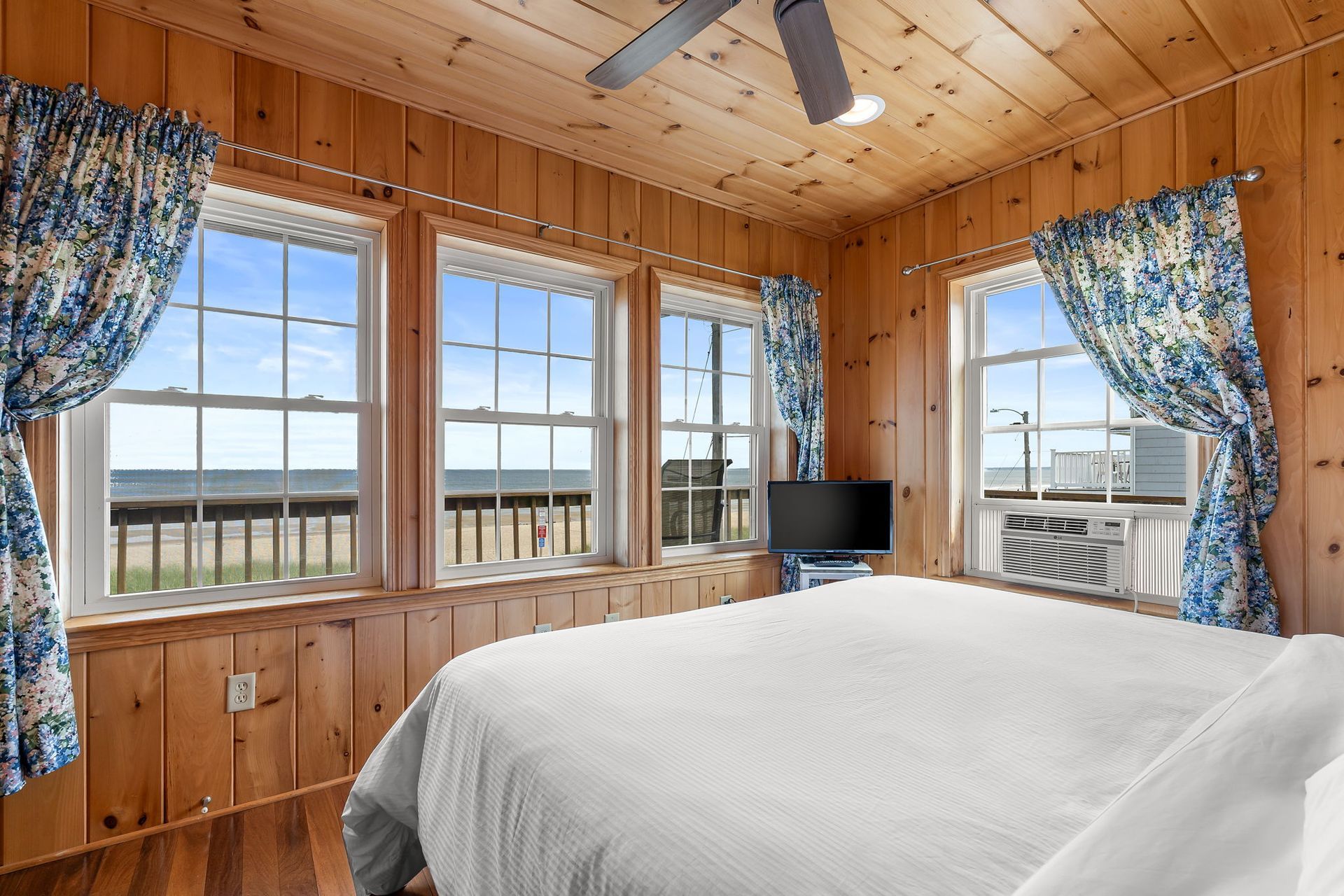 Bedroom with a beach view, wood-paneled walls, large windows, blue patterned curtains, and a white bedspread.