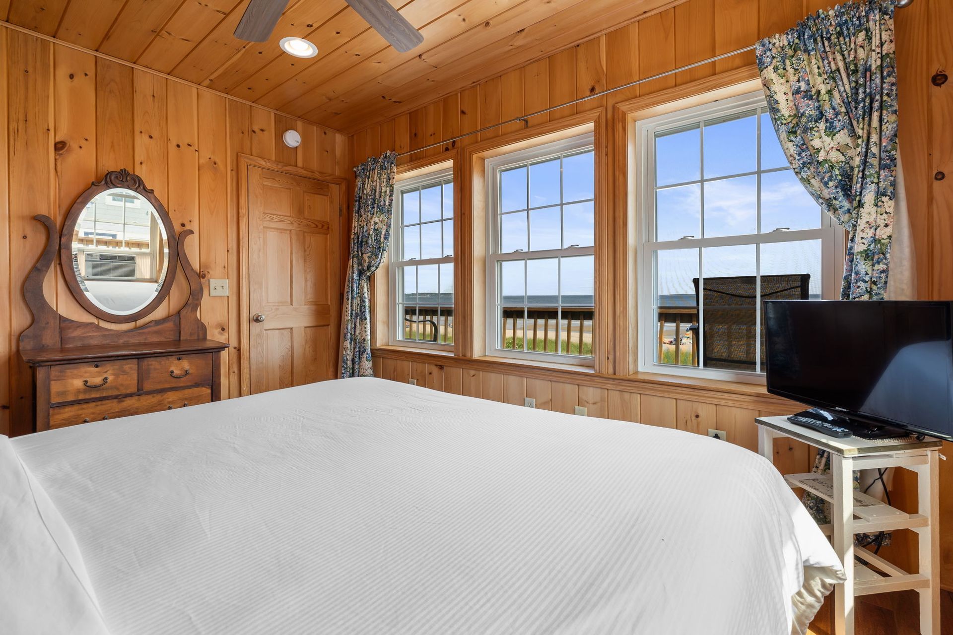 Bedroom with wood paneling, bed, windows, dresser, and TV. Blue curtains, ocean view.