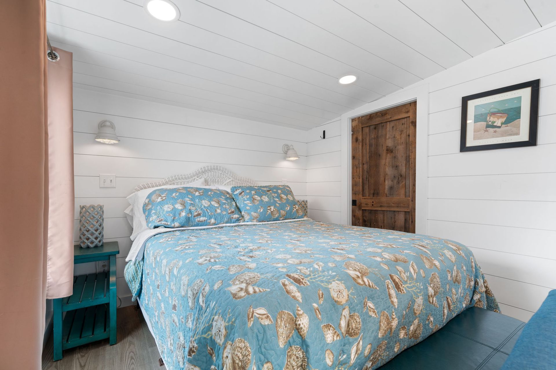 Bedroom with a blue floral bedspread, white shiplap walls, turquoise nightstand, and a wooden door.