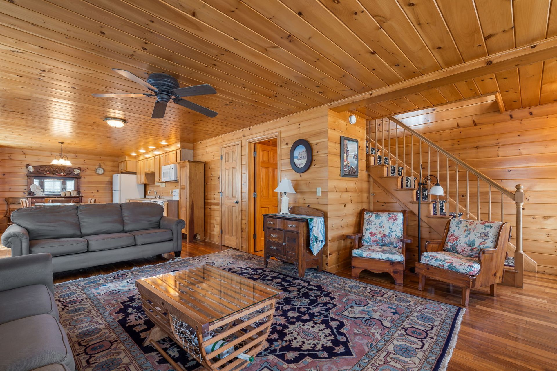 Living room with wood paneling, grey sofa, patterned rug, wooden chairs, and staircase.