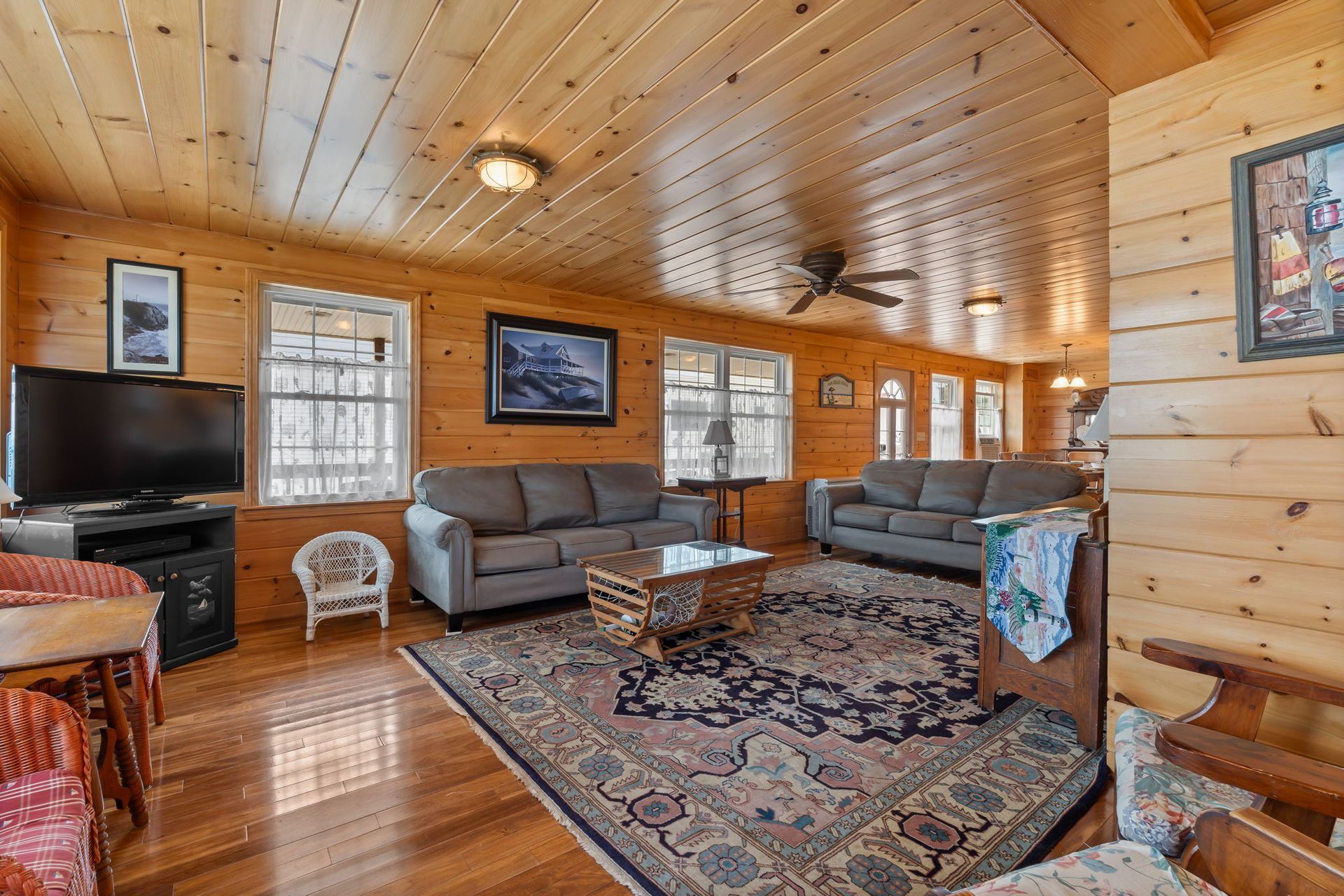 Cozy living room with wood paneling, two gray sofas, patterned rug, TV, and windows, typical of a cabin.