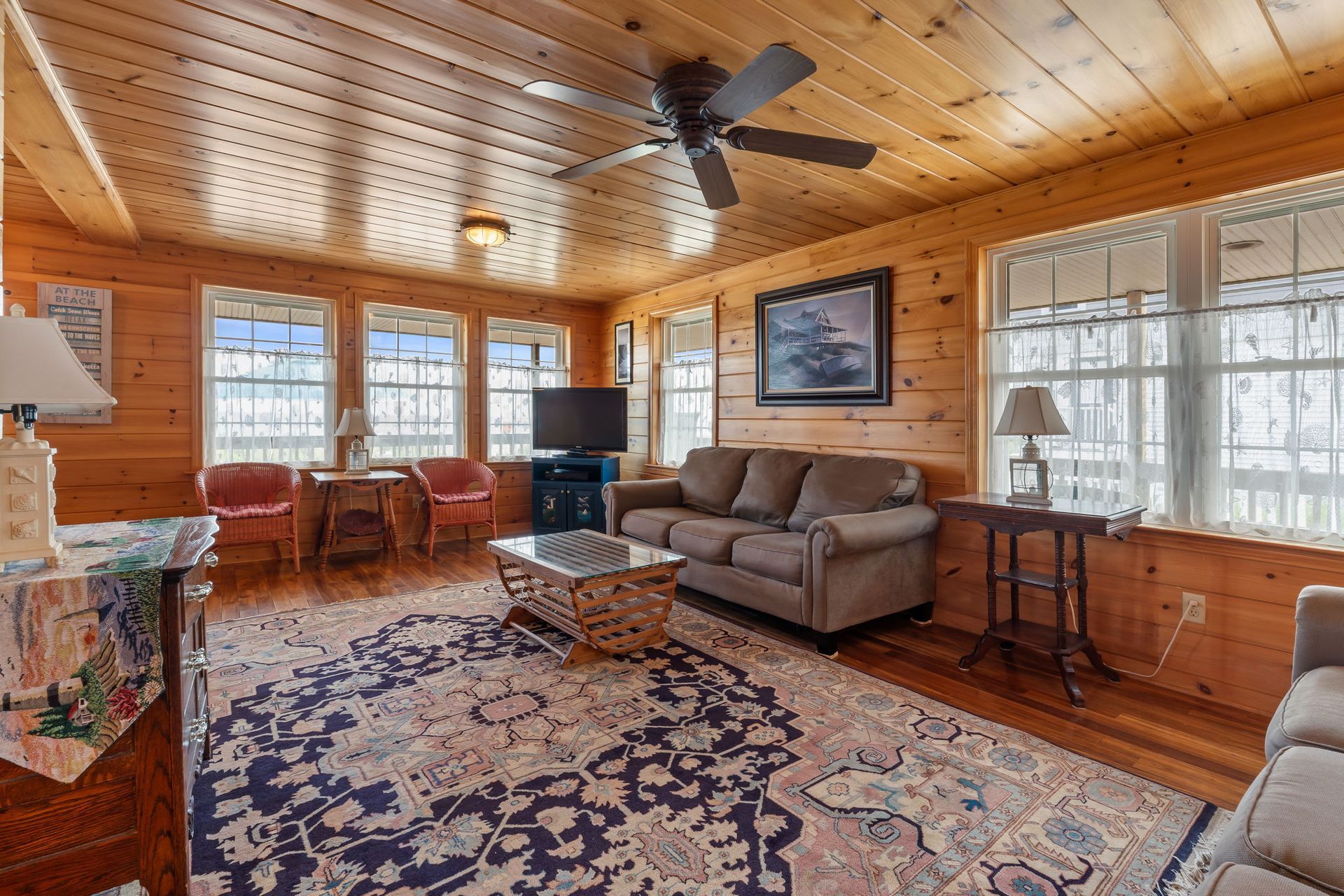 Cozy cabin living room with wood paneling, windows, sofa, rug, and ceiling fan.