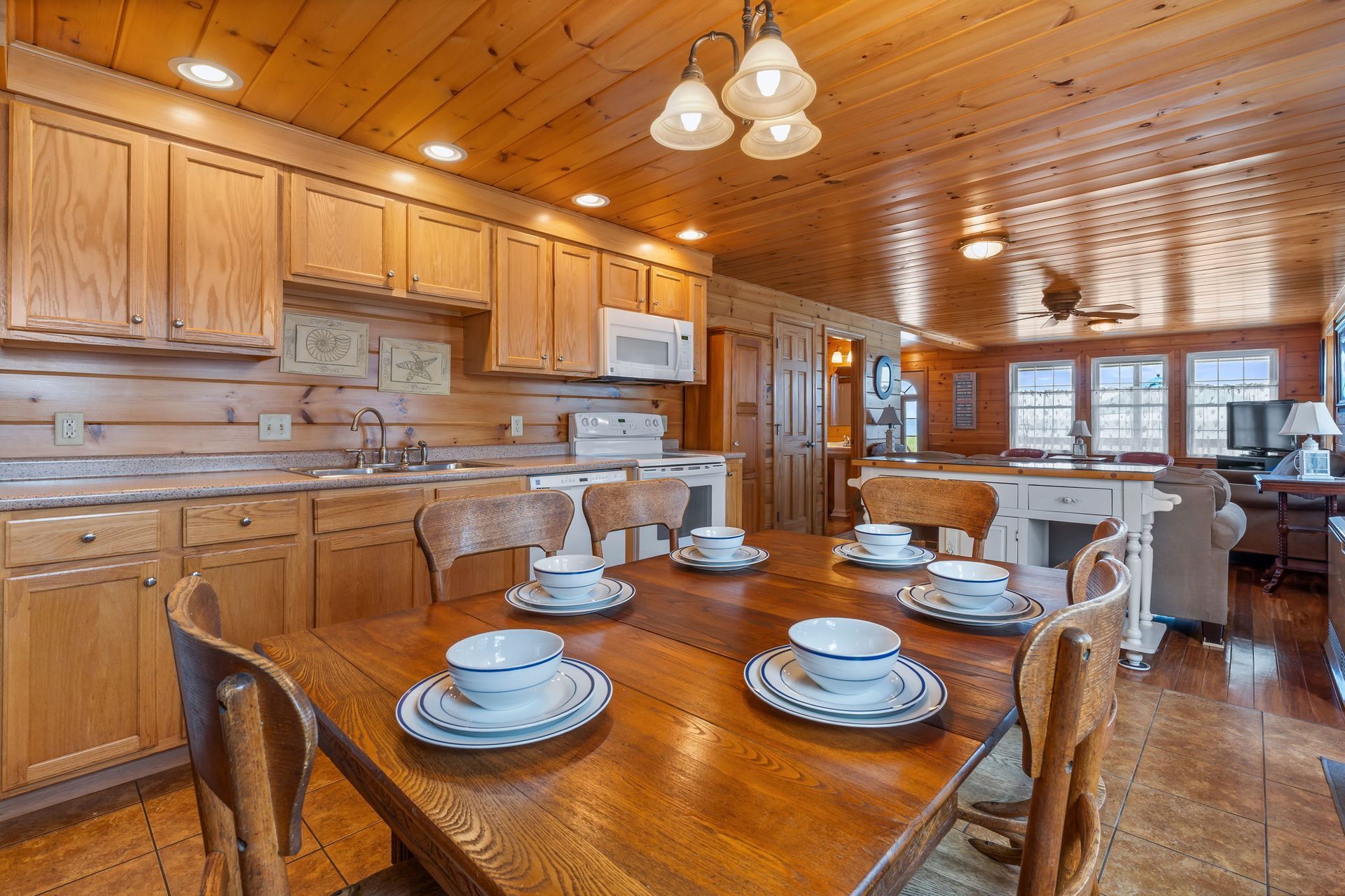 Wooden dining table set for six in a kitchen, with cabinets, appliances, and a view of a living area.
