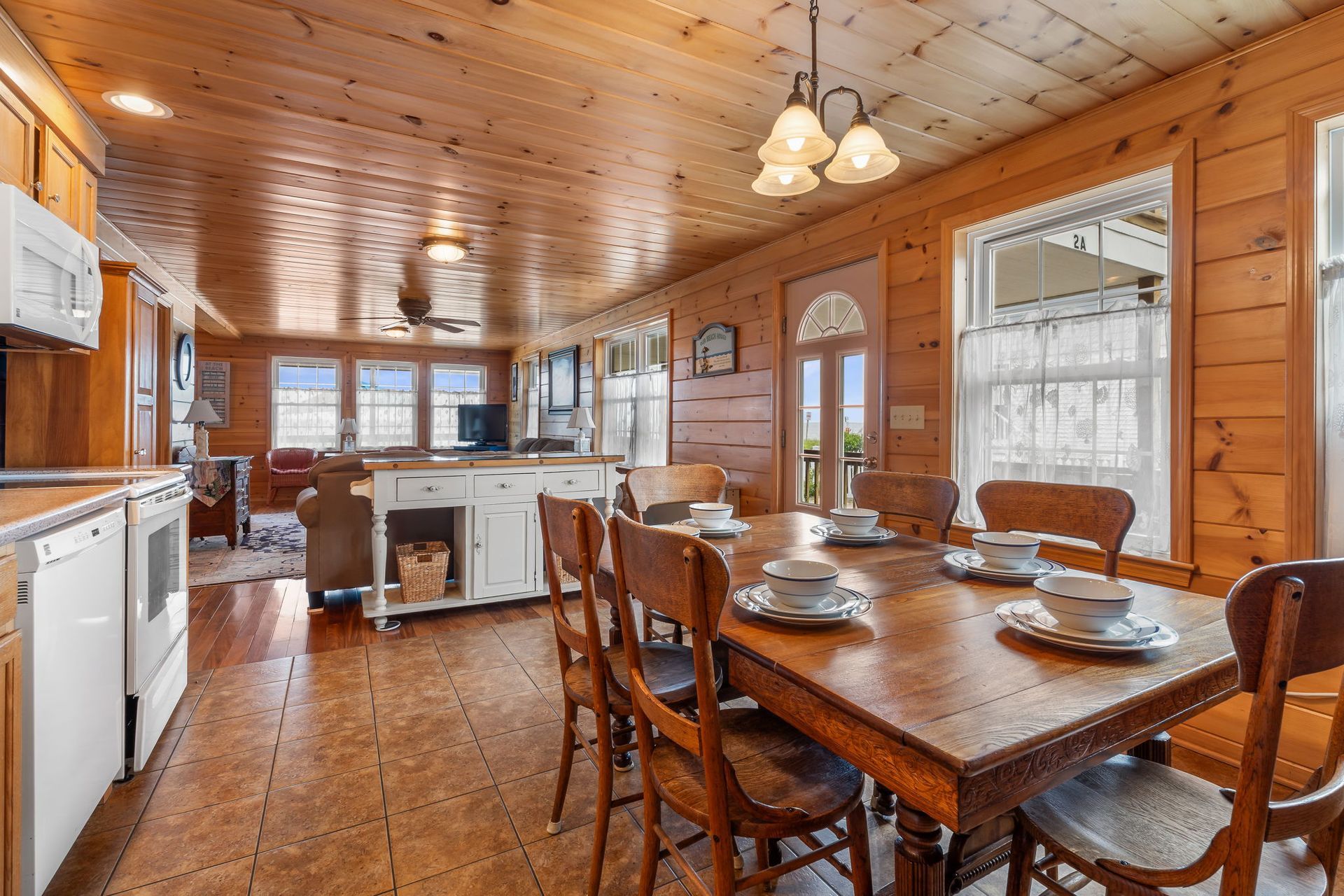 Wooden dining room with a table set for six; kitchen appliances on the left, windows on the right.