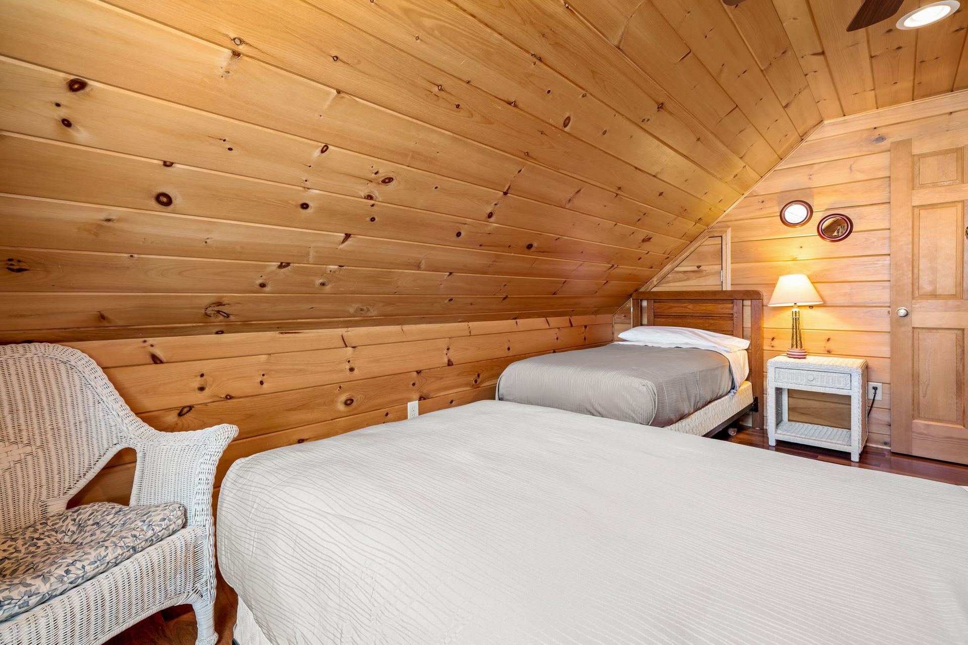 Cozy bedroom with wood paneling, two beds, a white wicker chair, and a nightstand with a lamp.