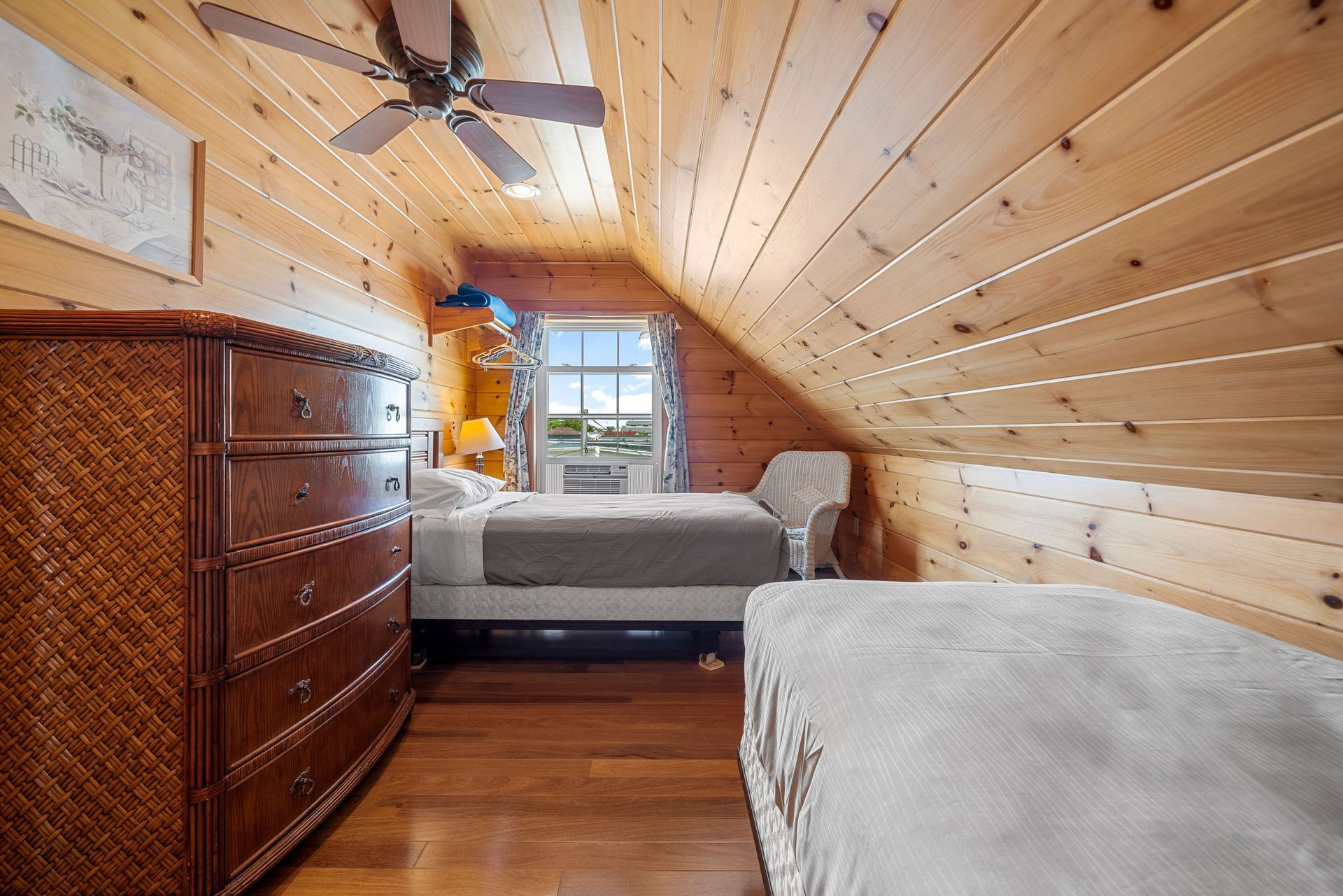 Cozy attic bedroom with wood paneling, bed, dresser, and bench, with a small window offering an outdoor view.