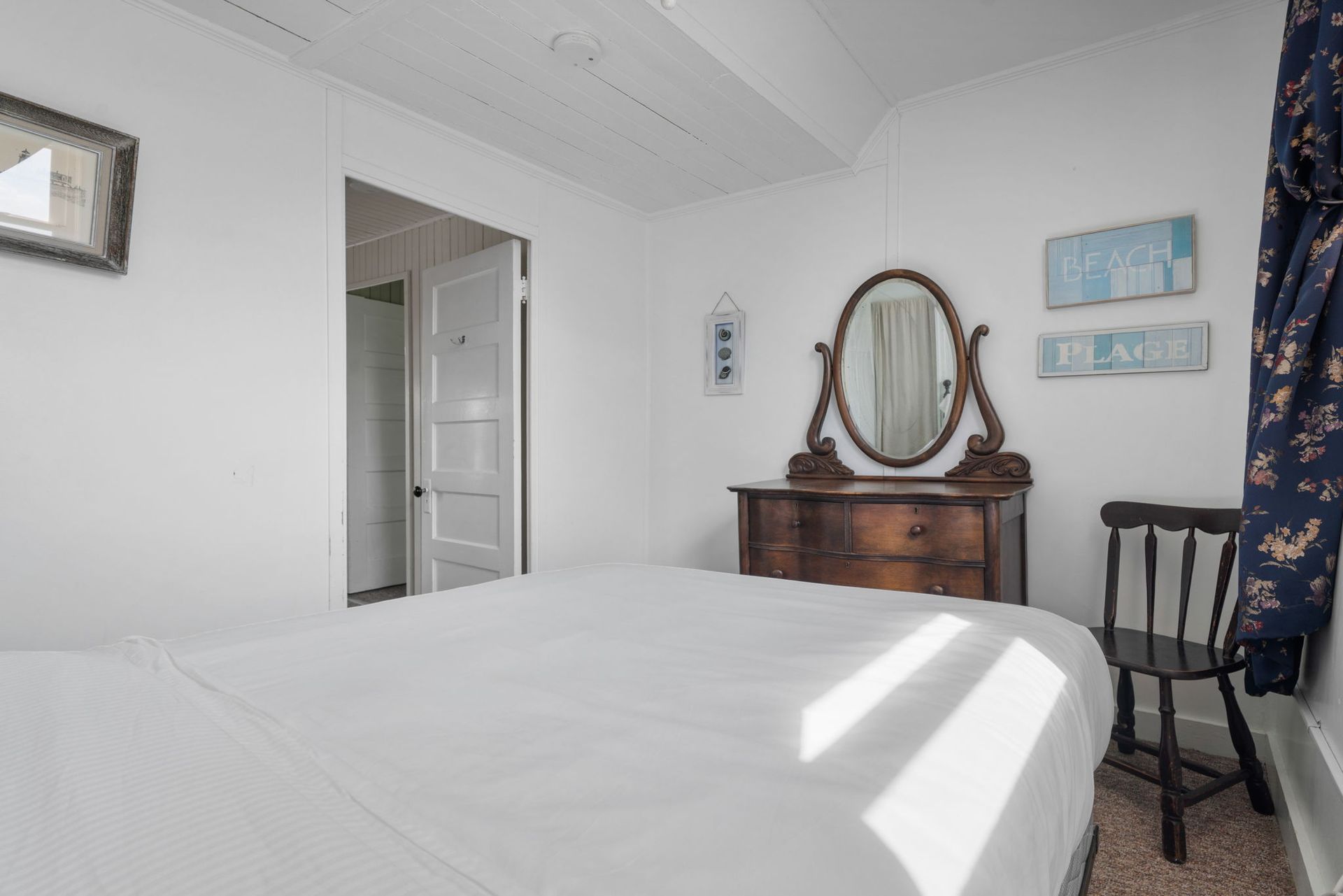 Bedroom with white walls, bed, dresser with mirror, and chair. Light streams in.