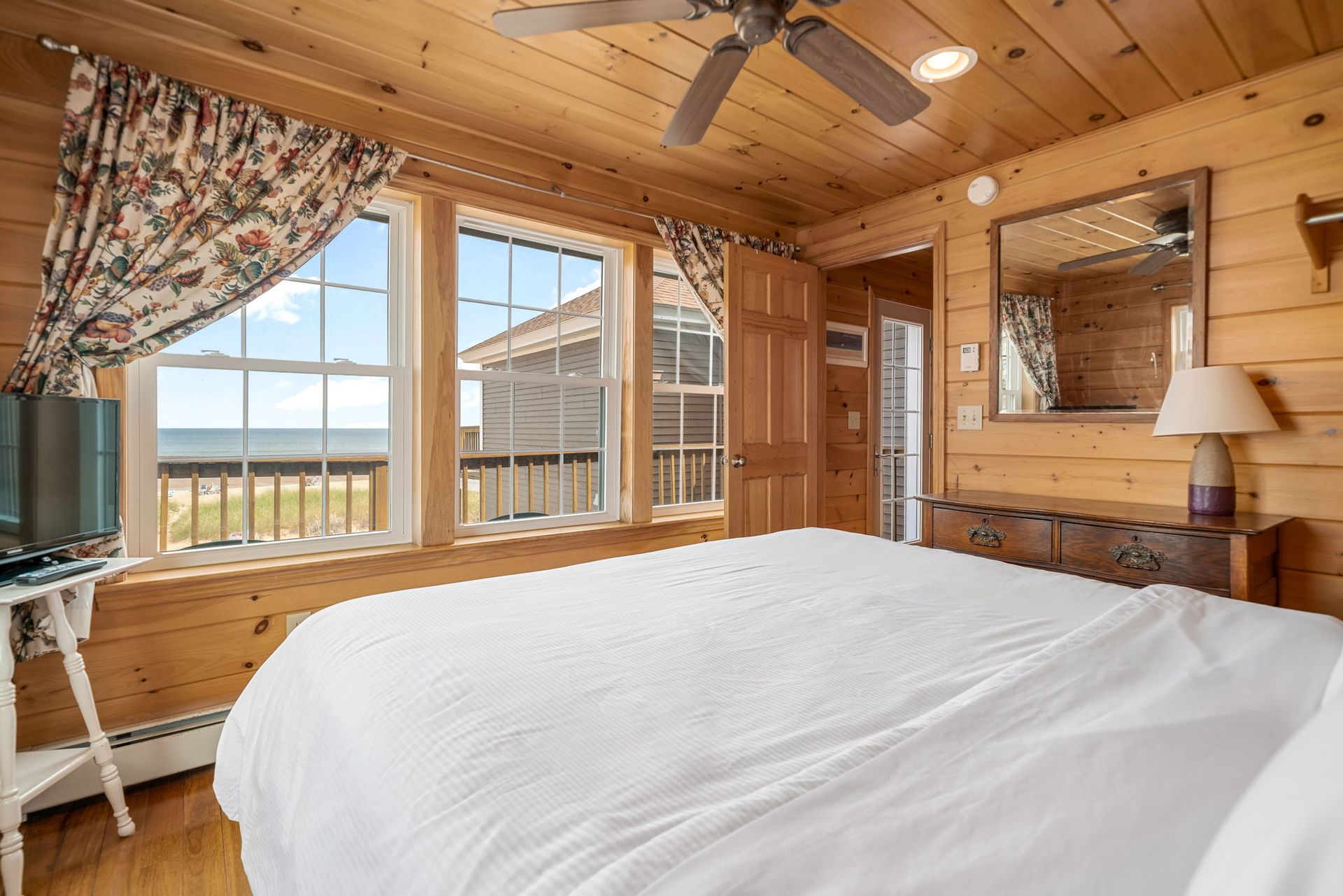 Bedroom with wood-paneled walls, white bed, ocean view, and floral curtains.
