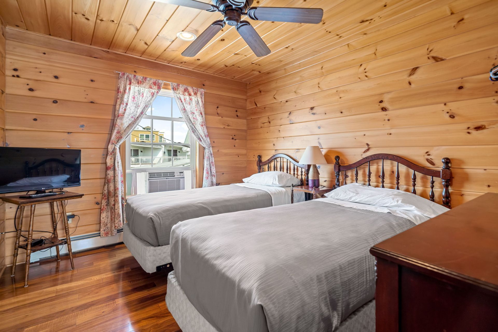 Bedroom with twin beds, wood-paneled walls, window with curtains, TV, and a ceiling fan.