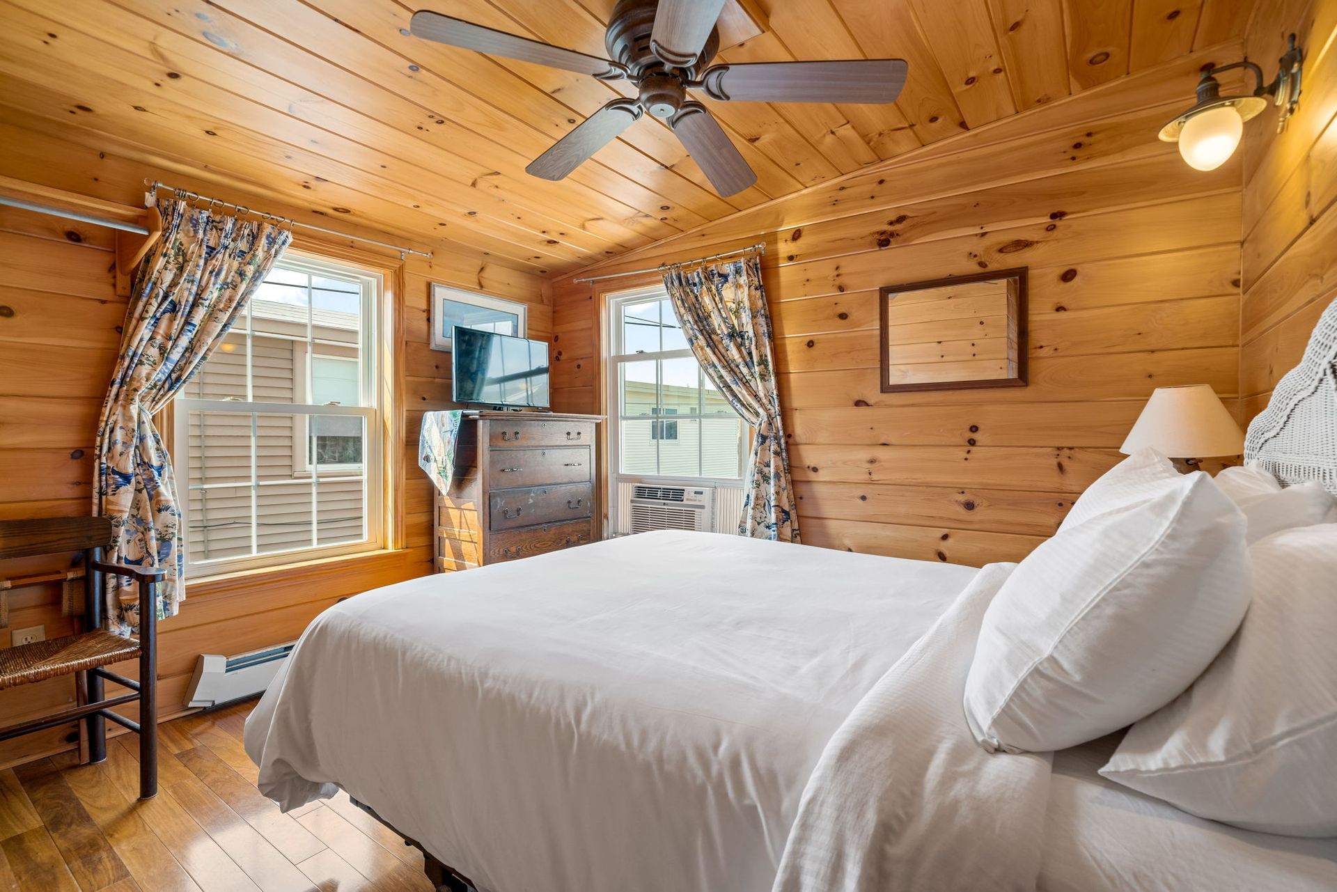 Bedroom with wooden walls, queen bed, windows with curtains, and ceiling fan.