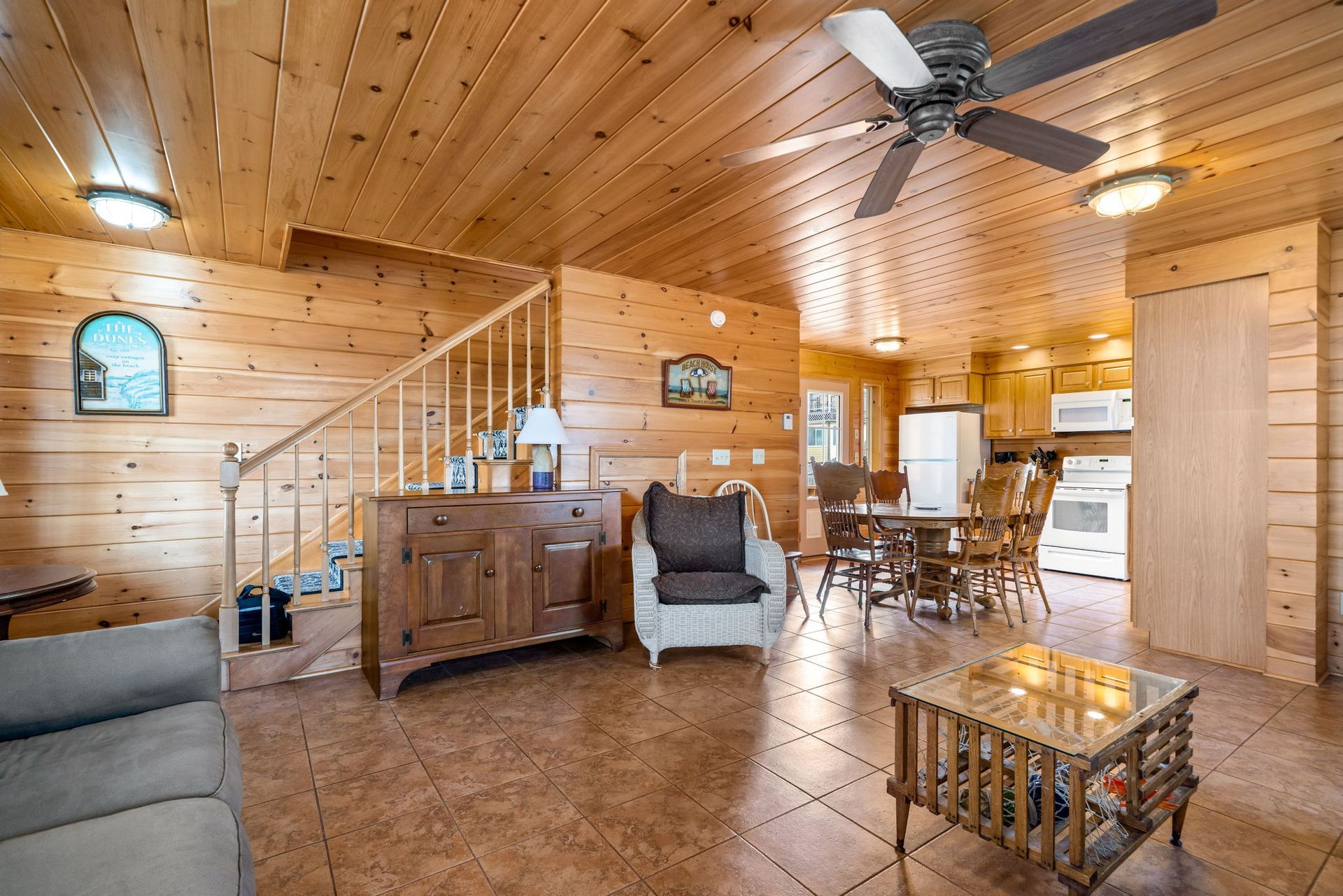 Cozy cabin interior: wooden walls/ceiling, staircase, living room, kitchen, brown flooring, dining table, ceiling fan.