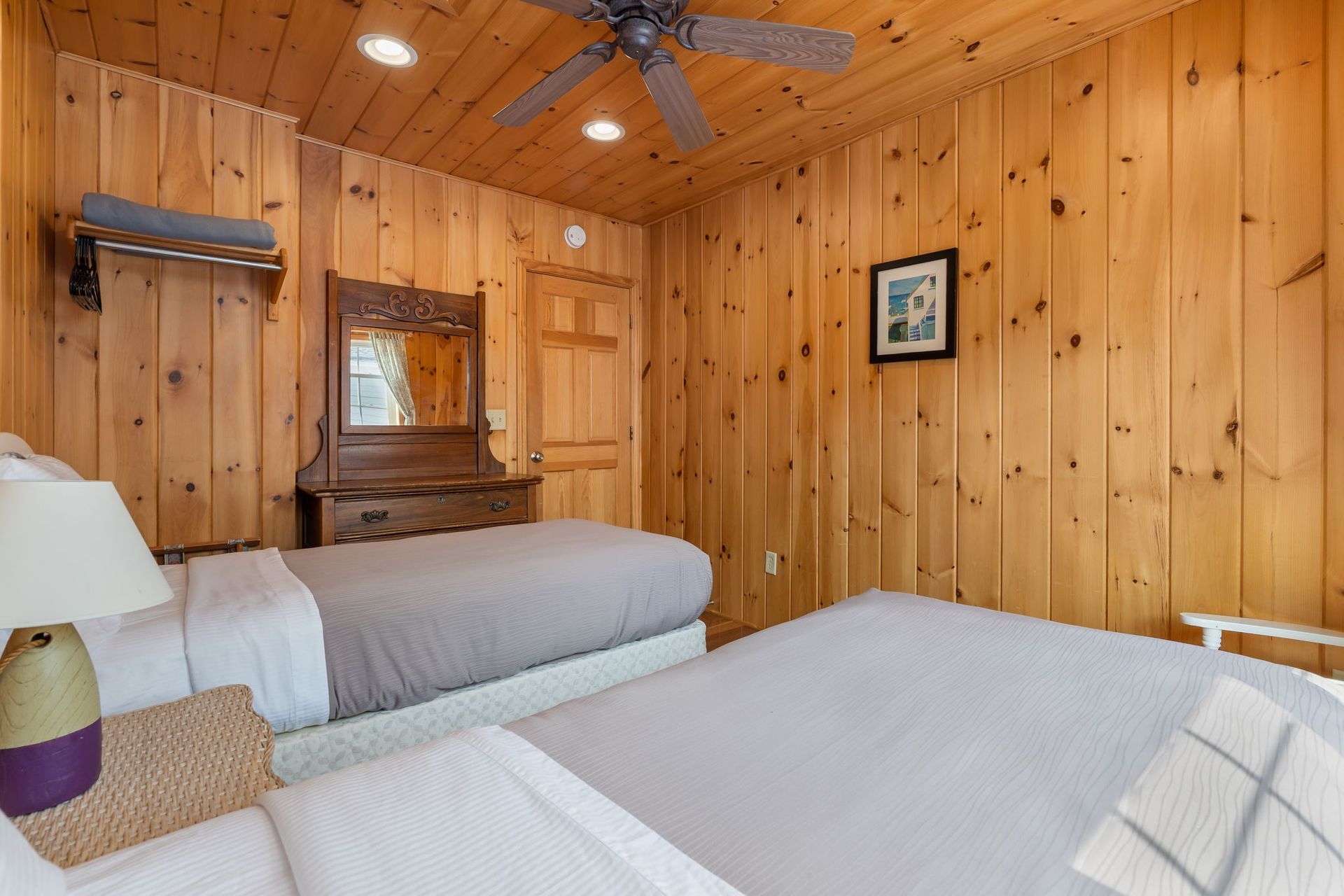 Bedroom with two beds, wood-paneled walls and ceiling.  A wooden dresser with a mirror and a small painting.