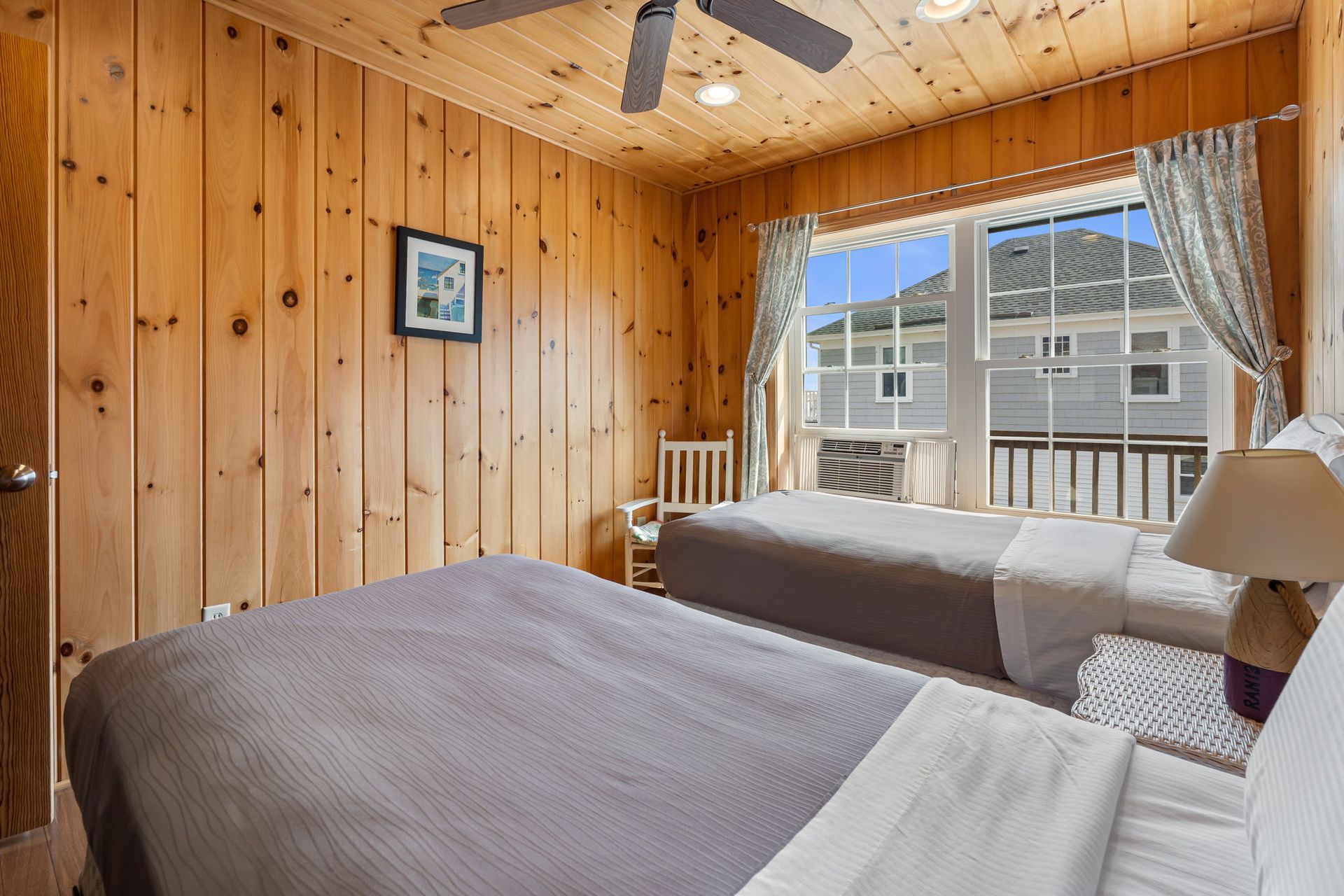 Bedroom with two twin beds, wood-paneled walls, window with view, and ceiling fan.