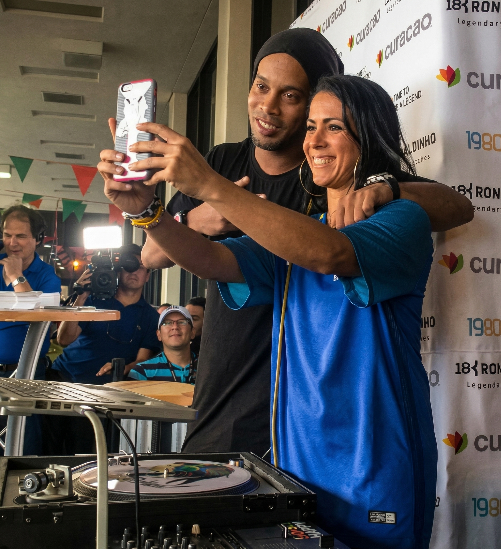 Duas pessoas sorrindo e tirando uma selfie na cabine do DJ durante um evento em local fechado.