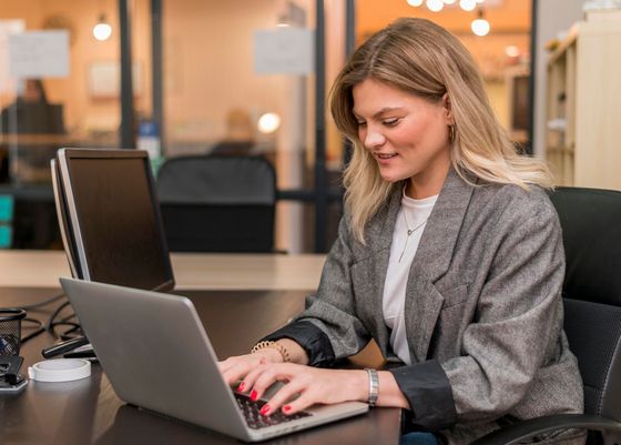 Woman working on laptop in office, typing with a smile, wearing gray blazer.