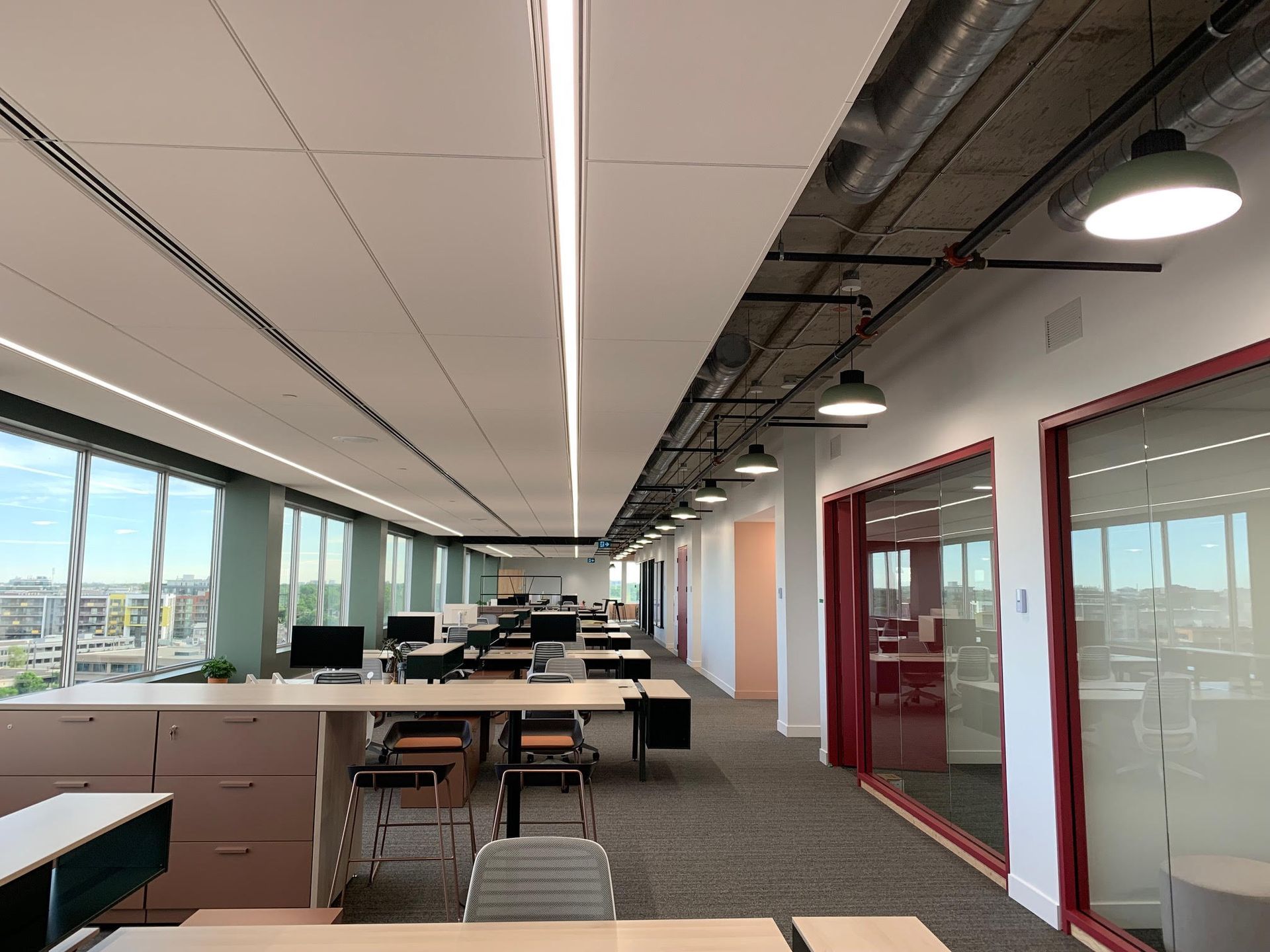 Espace de bureau moderne et lumineux avec de longs bureaux, des ordinateurs et des pièces aux parois vitrées le long d'un couloir.