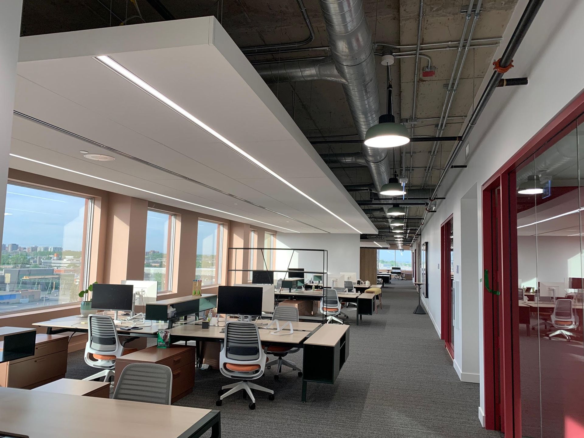 Bureau moderne avec bureaux, chaises et grandes fenêtres. Rangées de postes de travail avec ordinateurs, couleurs neutres et plafond apparent.