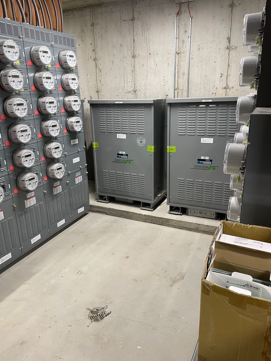 Local électrique avec panneau de compteurs, deux transformateurs gris et murs en béton.