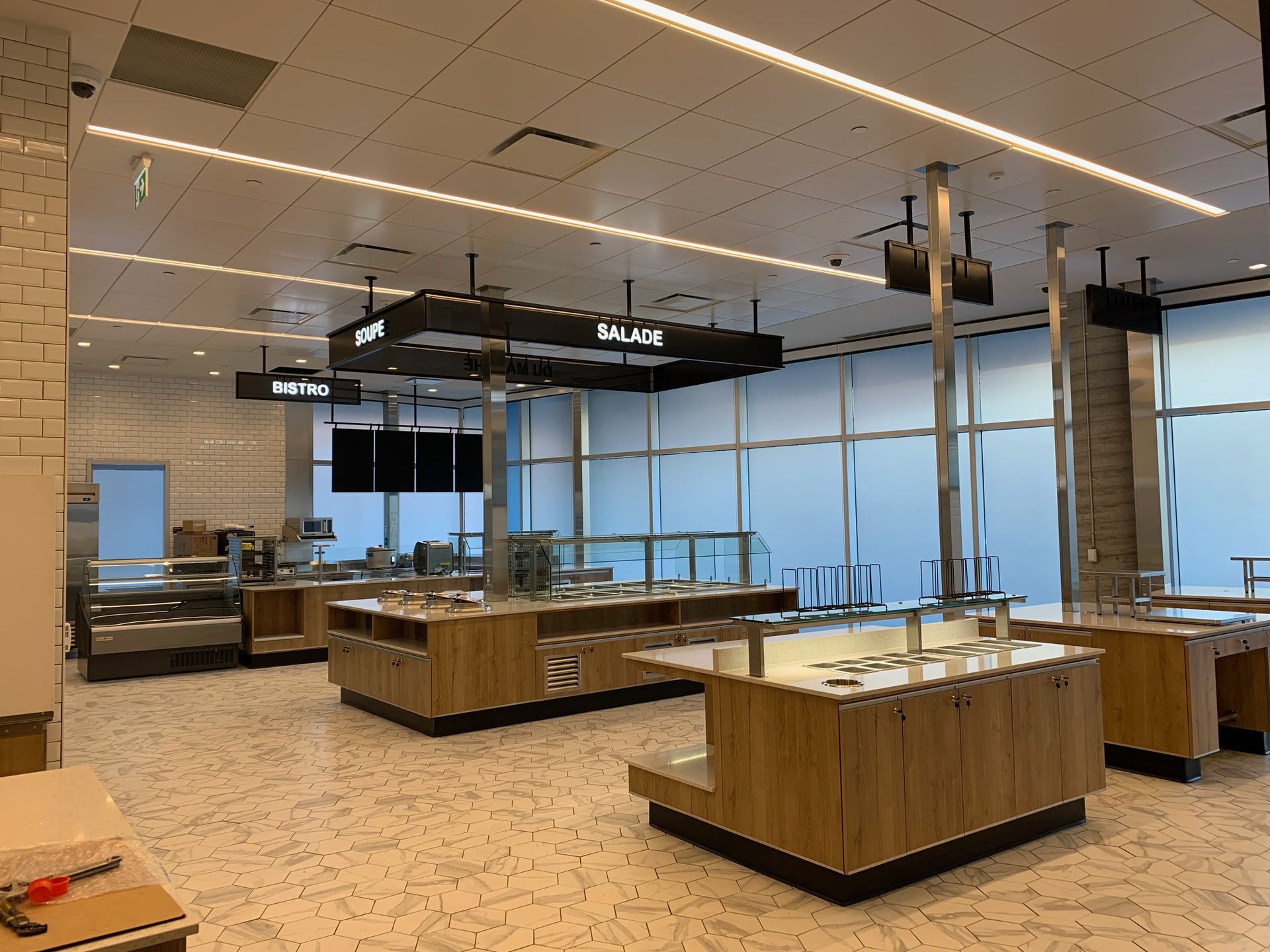 Cafétéria vide avec des stands de nourriture en bois, un éclairage au plafond et des panneaux indiquant « Salades » et « Soupe ».
