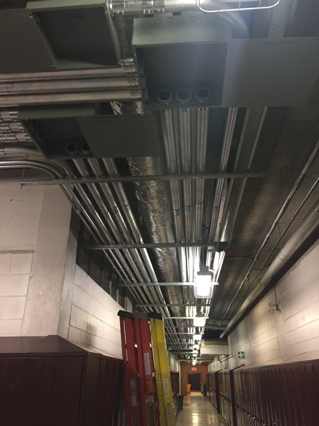 Couloir avec gaines aériennes, conduits et échelle. Murs blancs, casiers et éclairage fluorescent.