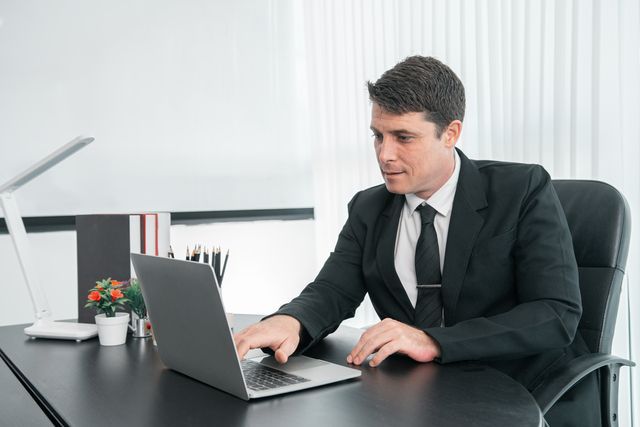 A man in a suit and tie is sitting at a desk using a laptop computer.