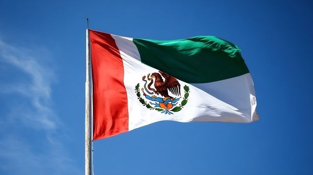 A mexican flag is waving in the wind against a blue sky.