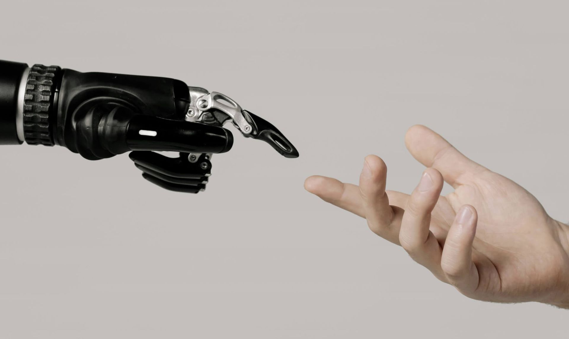 Uma mão robótica preta estendendo-se em direção a uma mão humana, ambas estendidas. Fundo cinza.