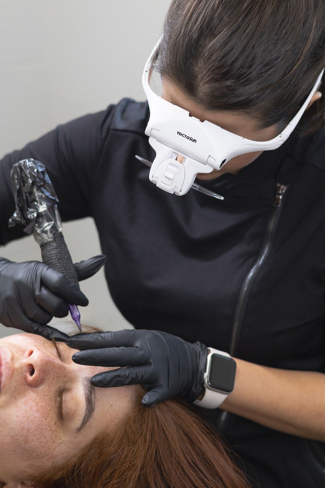 Woman getting eyebrow microblading, artist wearing magnification glasses and black gloves.