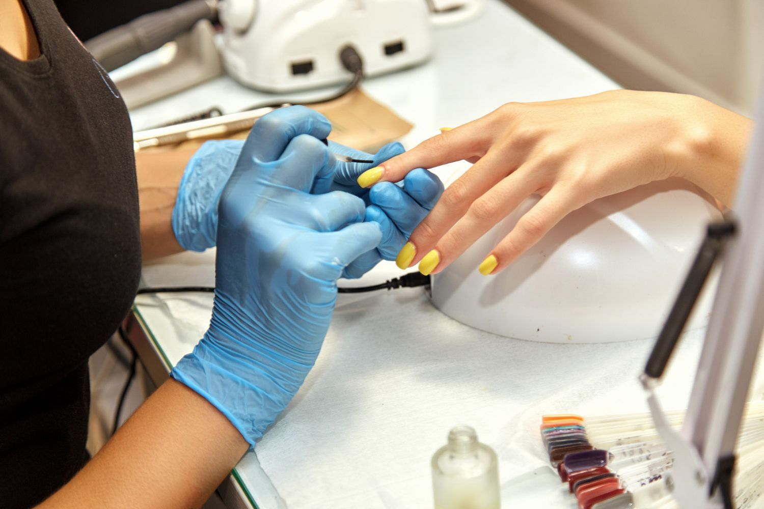 Manicurist filing a client's nails with a tool, client's hand with yellow polish on a white table.