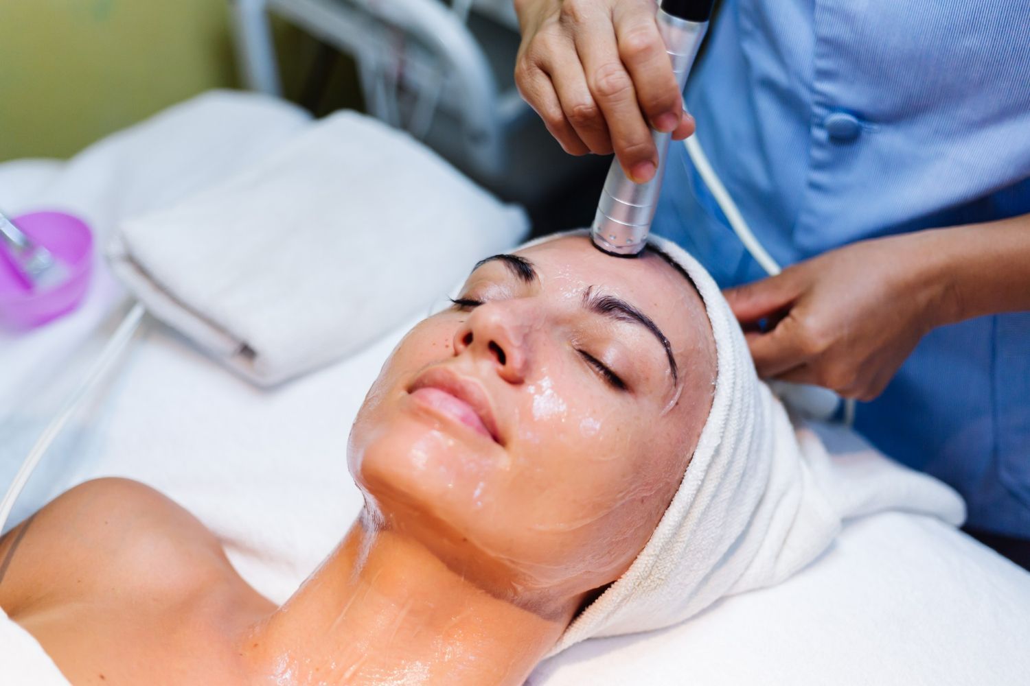 Woman receiving a facial treatment; technician holds tool to her forehead.