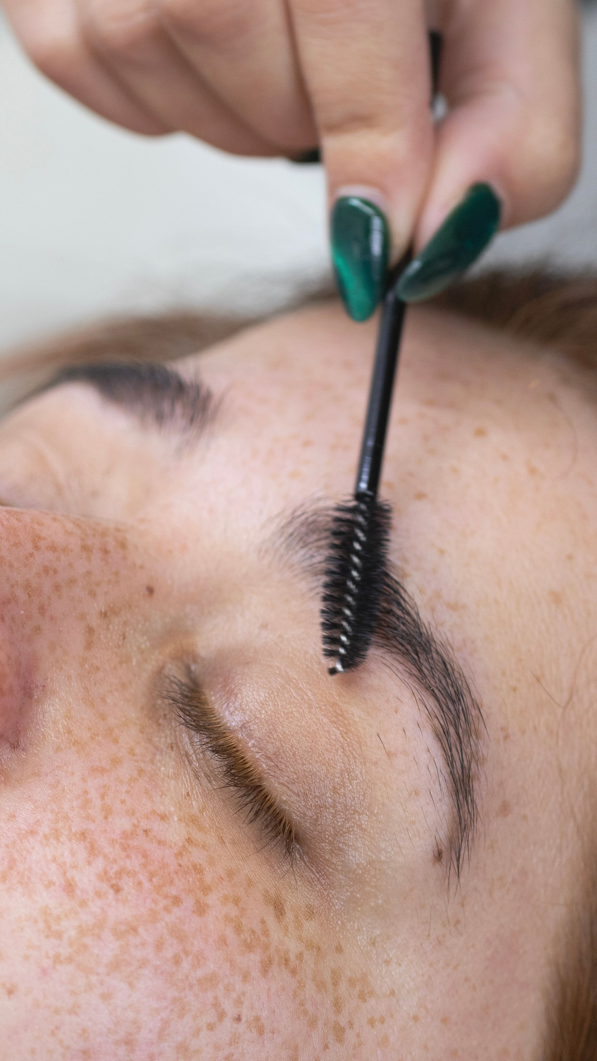 A woman with green nails is applying mascara to her eyebrows.