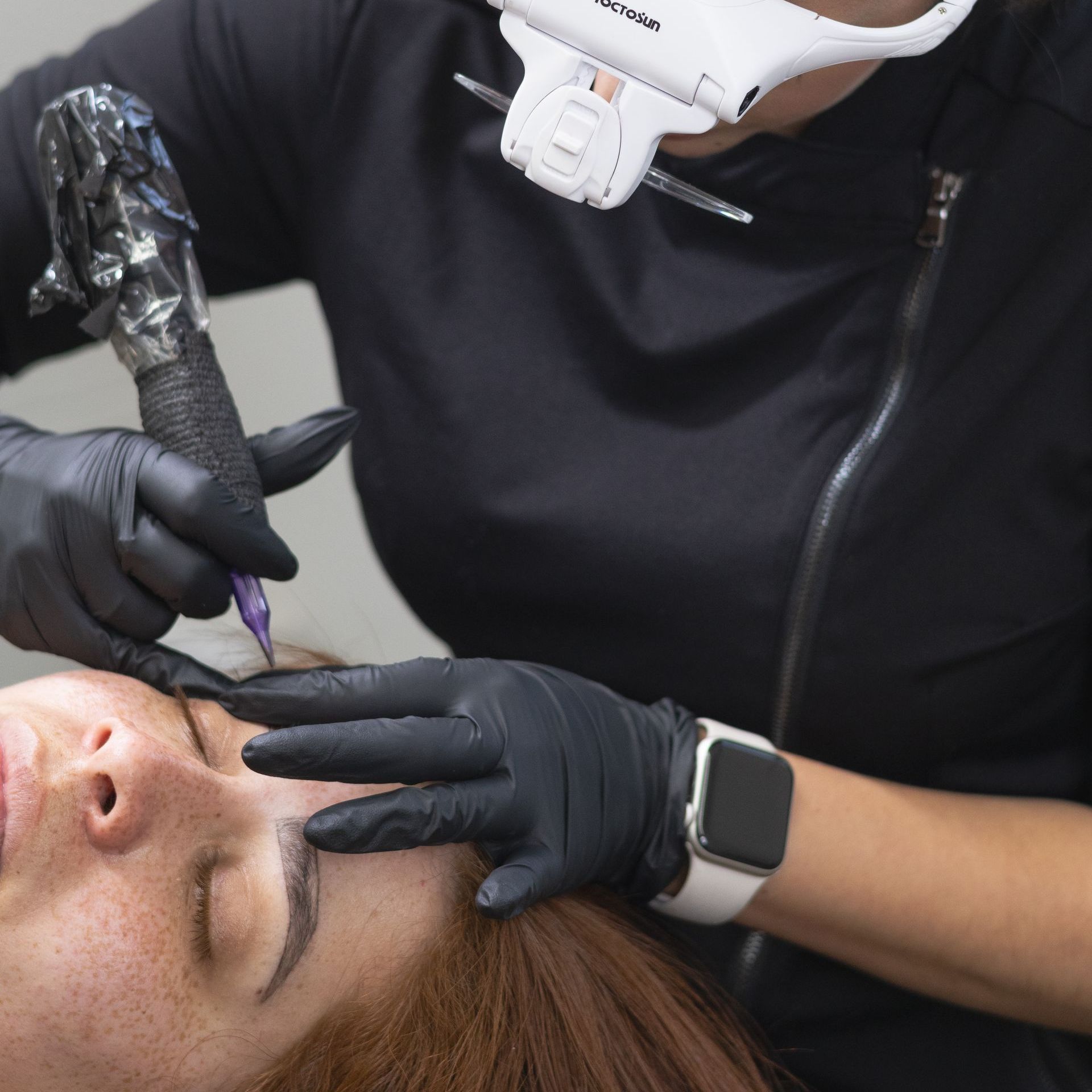 A woman wearing black gloves is getting a tattoo on her face