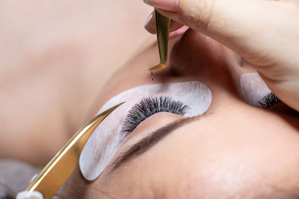 A person receiving eyelash extensions. A technician uses tweezers to apply an extension while the eye is taped.