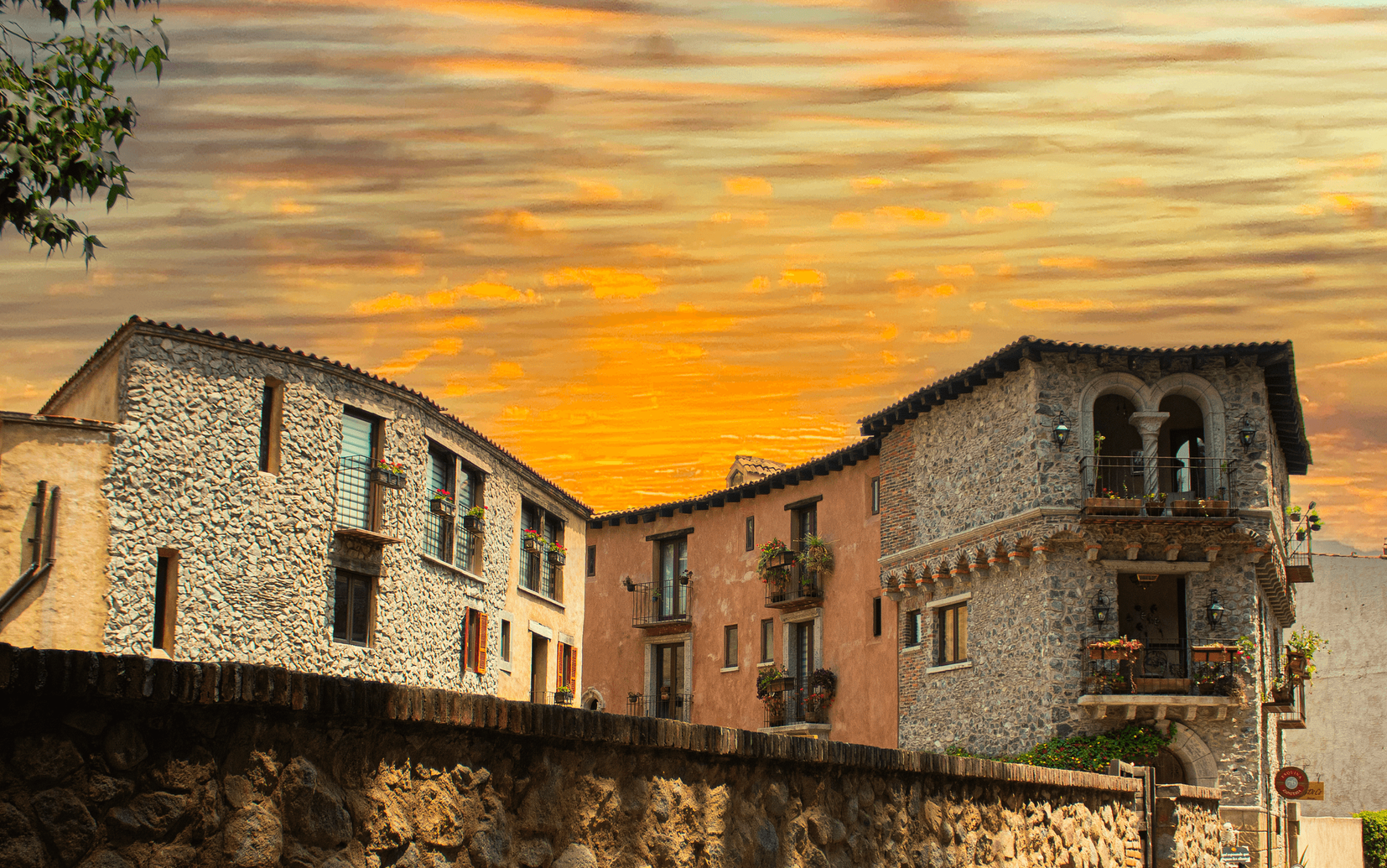 Stone buildings against a fiery sunset sky, with balconies and a stone wall in the foreground.