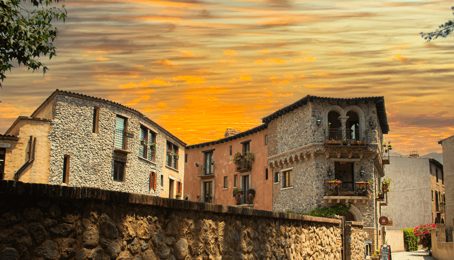 Stone buildings with balconies against a golden sunset sky. A stone wall is in the foreground.