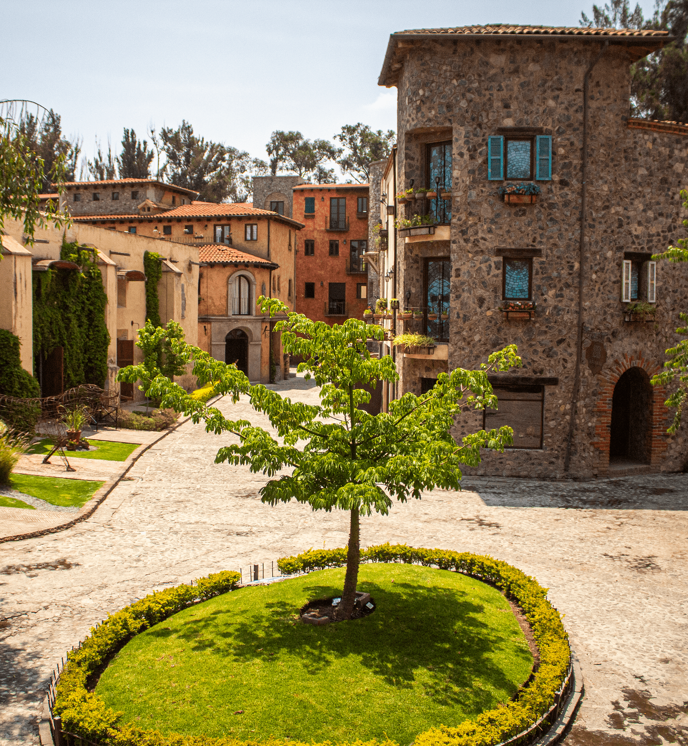 Courtyard of stone buildings, tree in a grass circle, cobblestone ground, blue shutters, sunny day.
